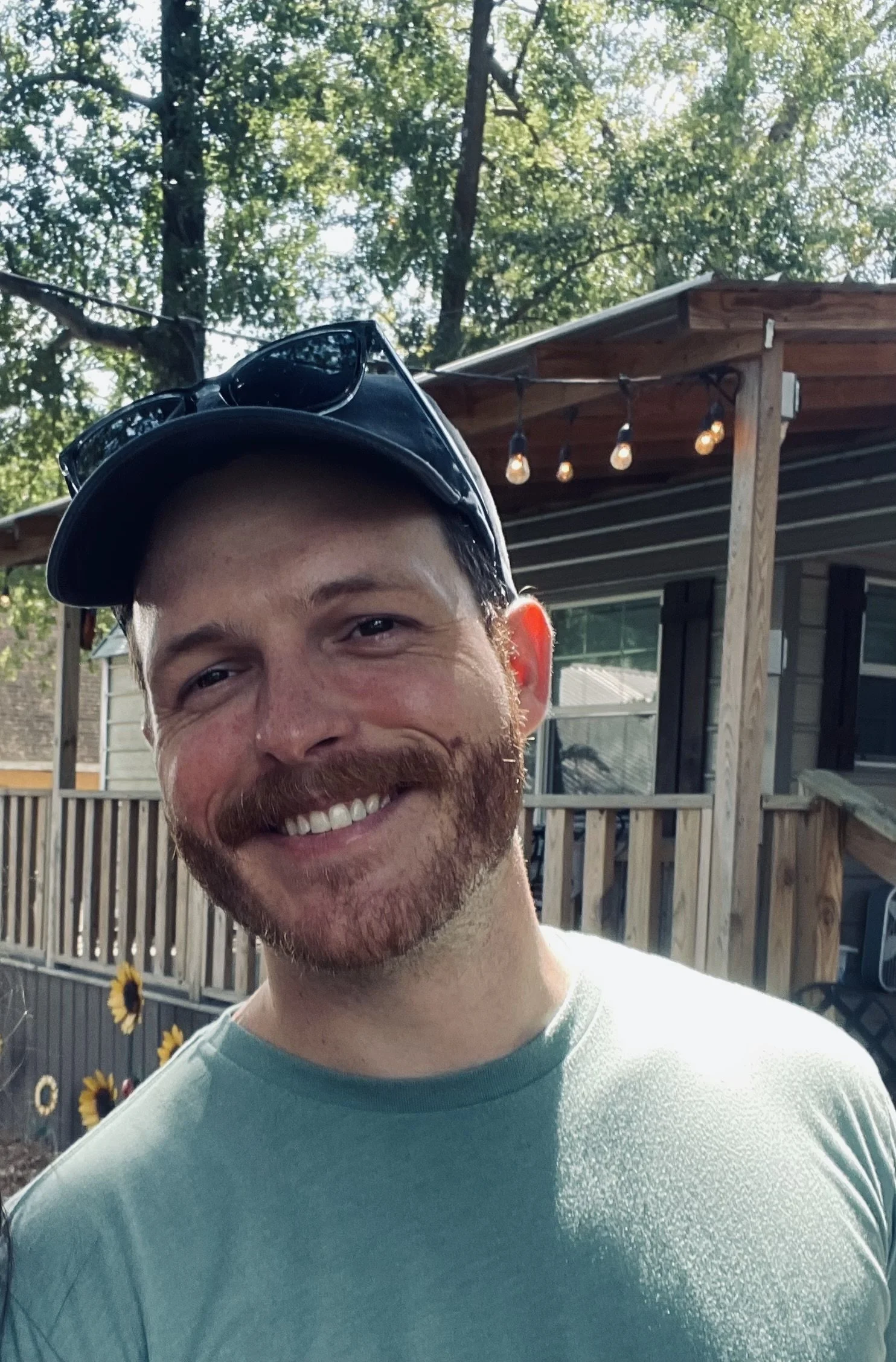 A smiling man with a beard wearing sunglasses on a cap, standing outdoors in a backyard with trees, a wooden deck, string lights, and sunflowers.