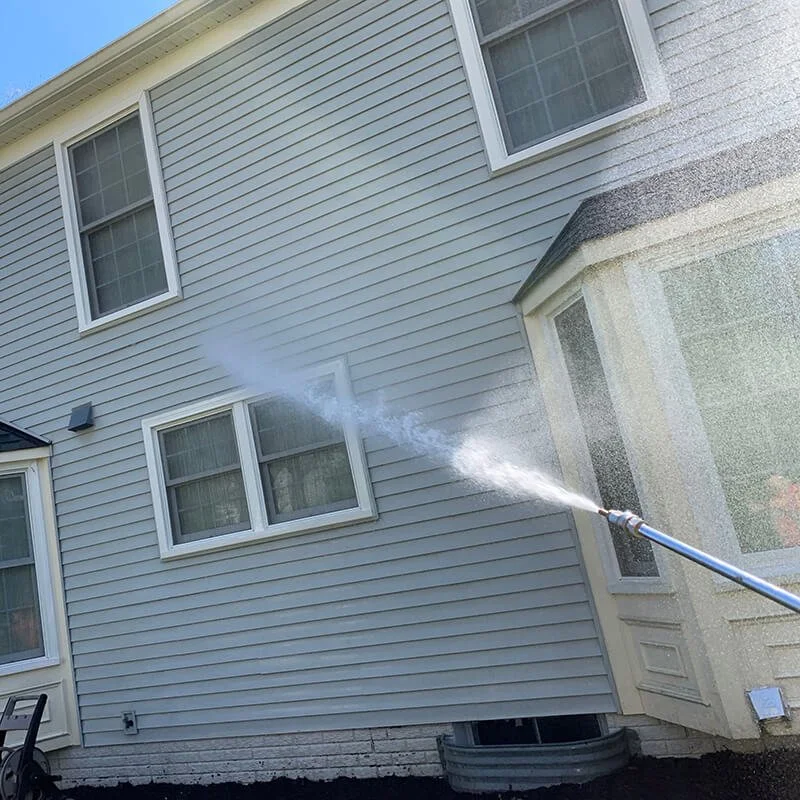 A high-pressure water hose cleaning the exterior wall of a house, which has beige siding and multiple windows.