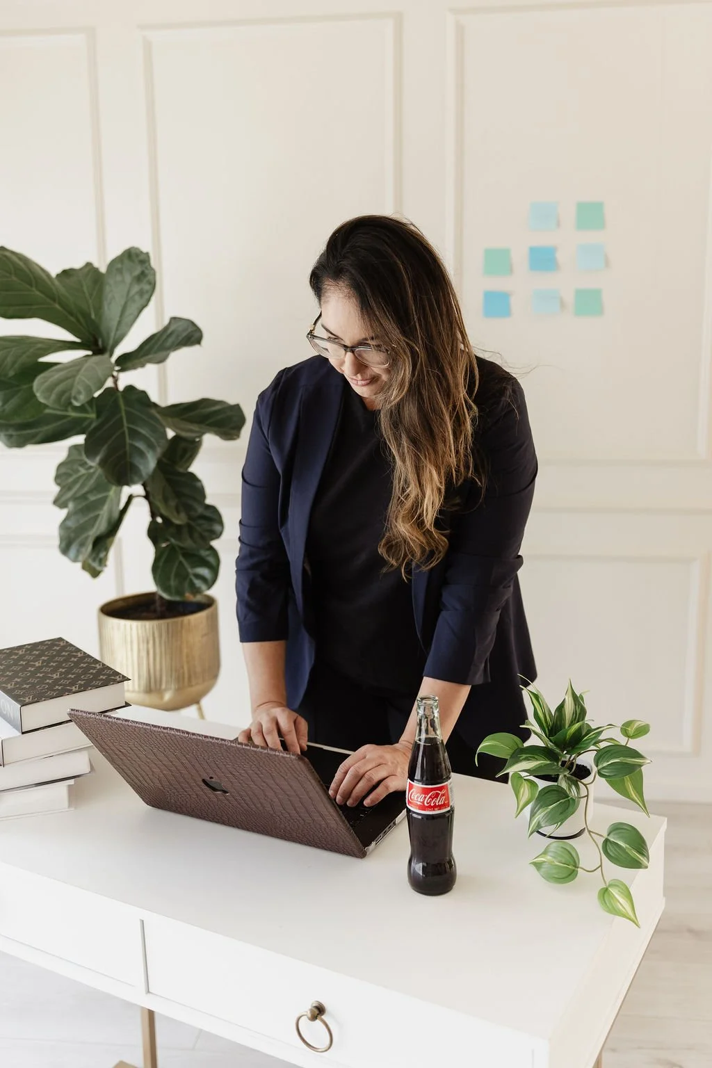 A woman in glasses and a navy blazer working on a laptop at a white desk with plants and a bottle of Coca-Cola.