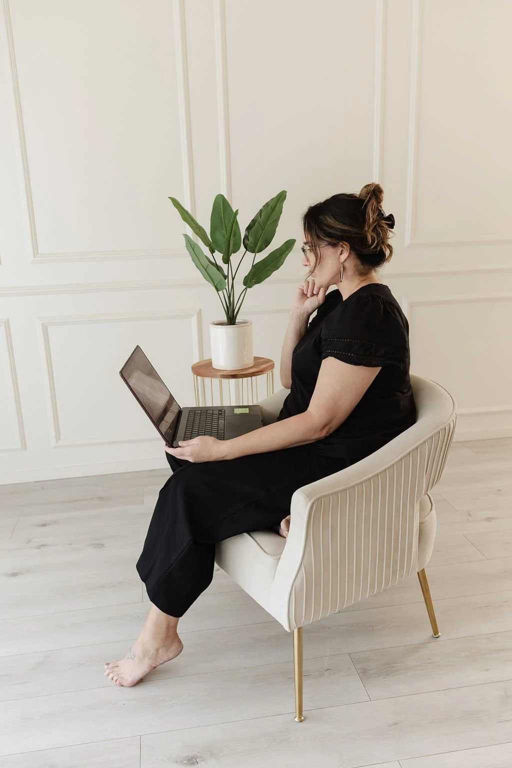 A woman with glasses sitting in a cream-colored chair, working on a laptop, with a potted plant nearby.