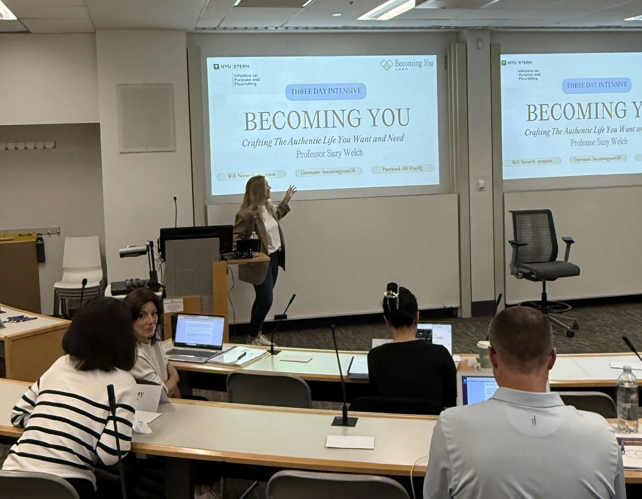 A woman standing at a podium in a classroom or conference room giving a presentation to attendees. The presentation slide behind her reads 'BEcoming YOU, Crafting The Authentic Life You Want and Need,' indicating a workshop or seminar on personal development.
