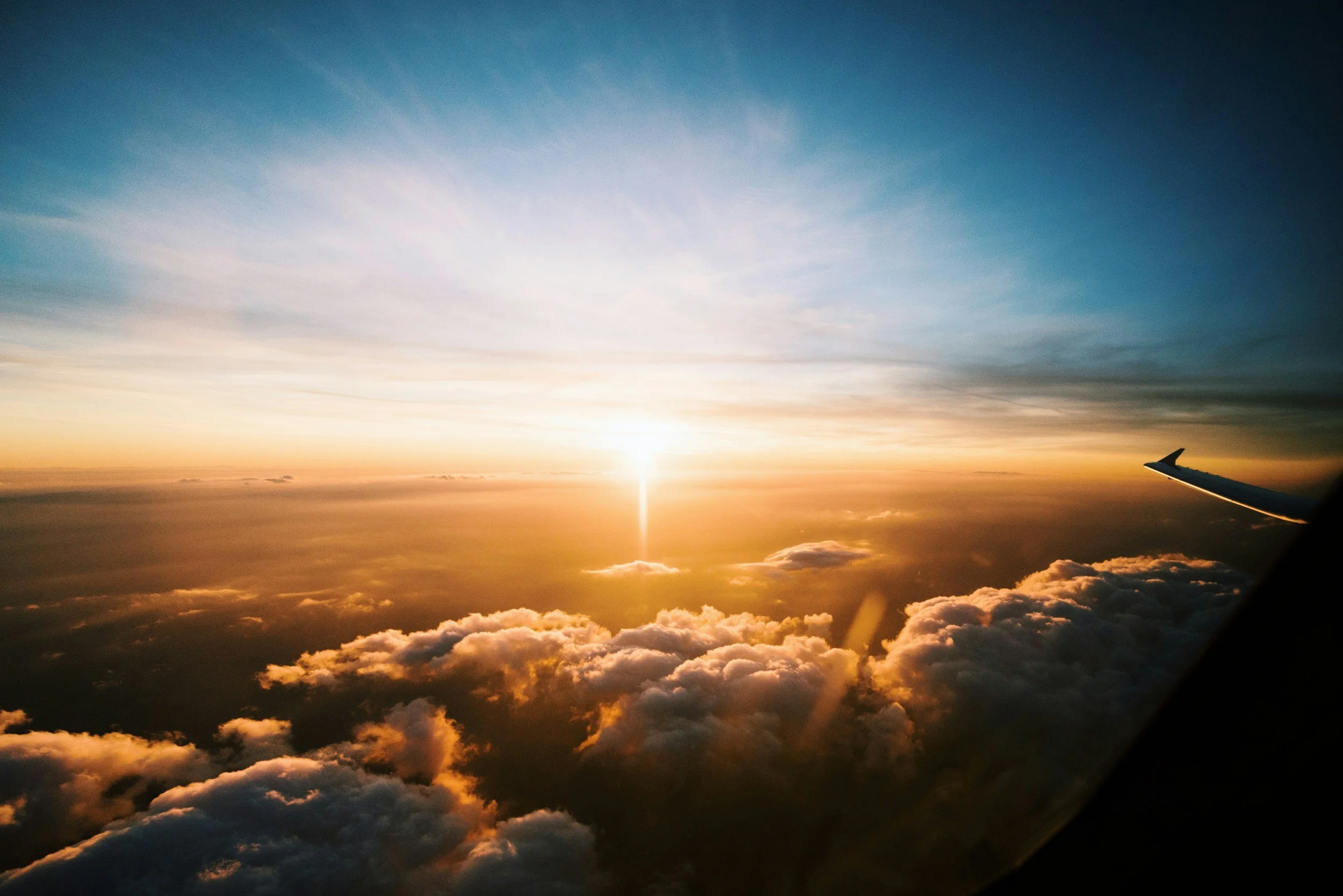 View from an airplane window showing a sunset or sunrise above clouds with part of the airplane wing visible on the right.