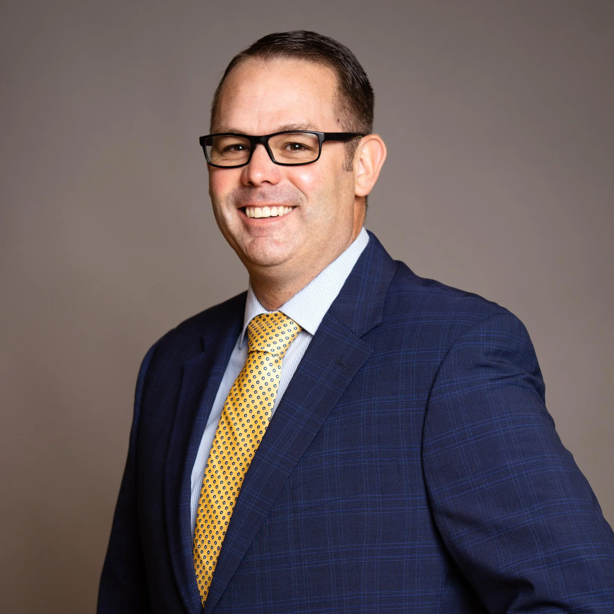 Professional portrait of a man in a blue suit, light blue shirt, yellow polka dot tie, wearing glasses, smiling against gray background.