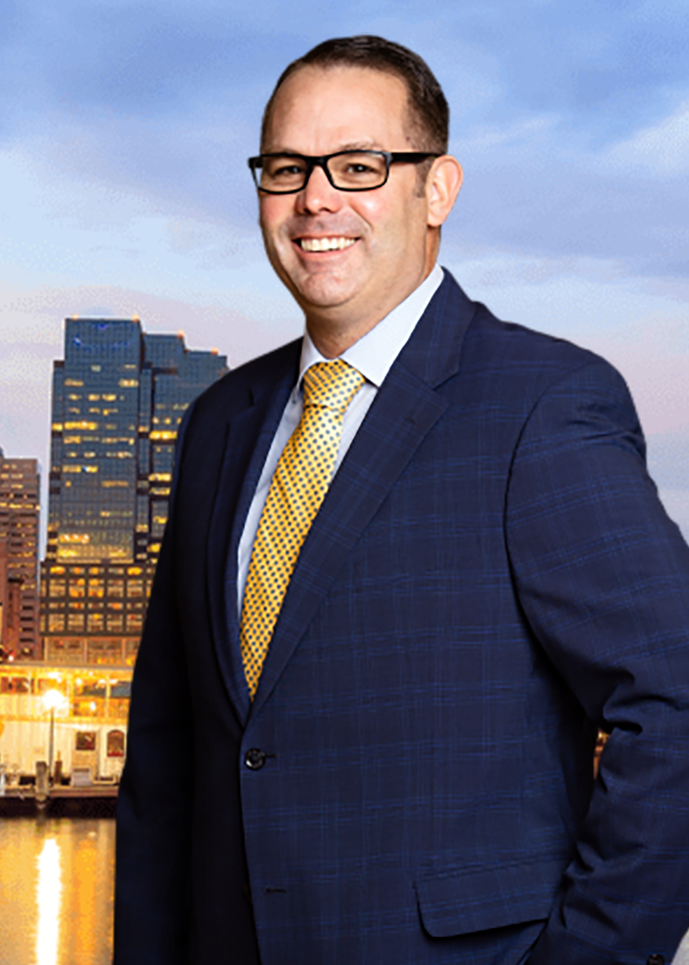 A man in a navy blue suit and yellow tie standing outdoors with city buildings and a river in the background, during dusk.