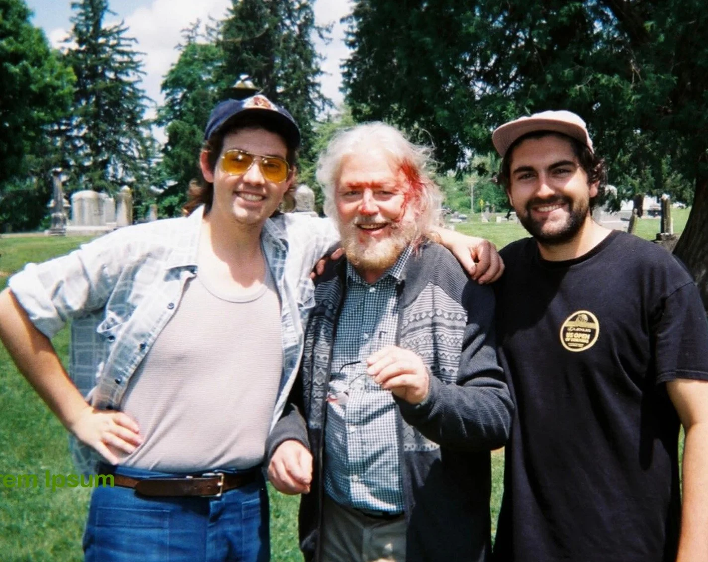 Three men standing together outdoors in a grassy area with trees and tombstones in the background. They are smiling and have their arms around each other.