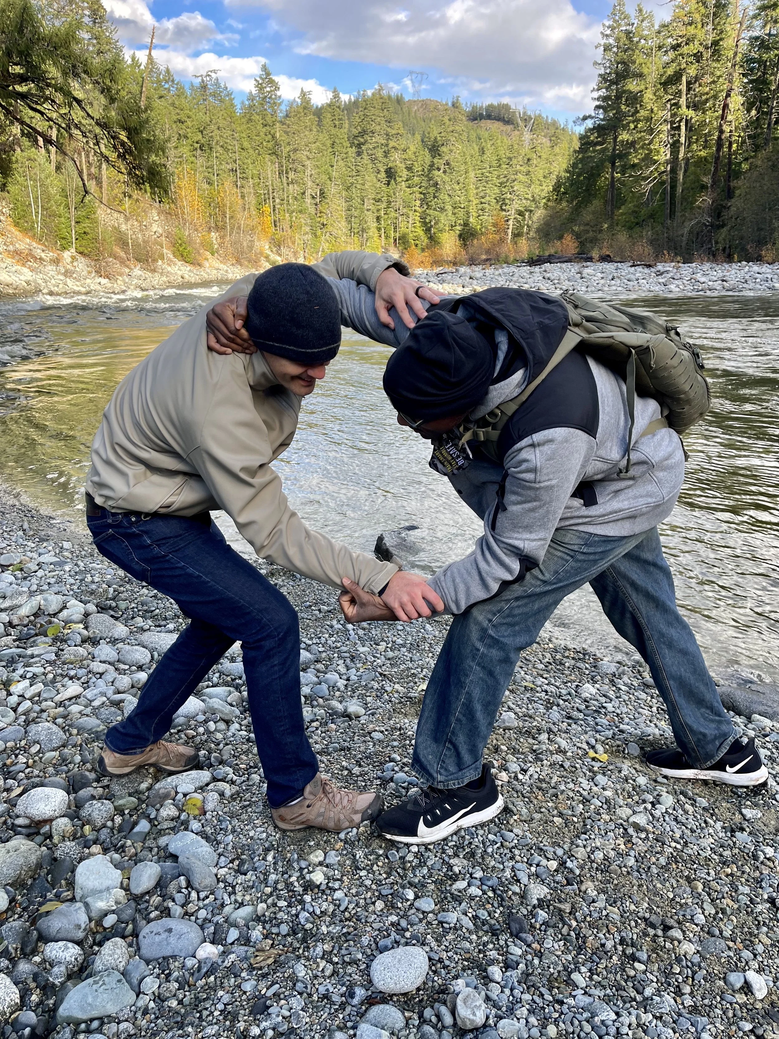 Two men struggling and fighting on a rocky riverbank, with a forested hillside in the background.