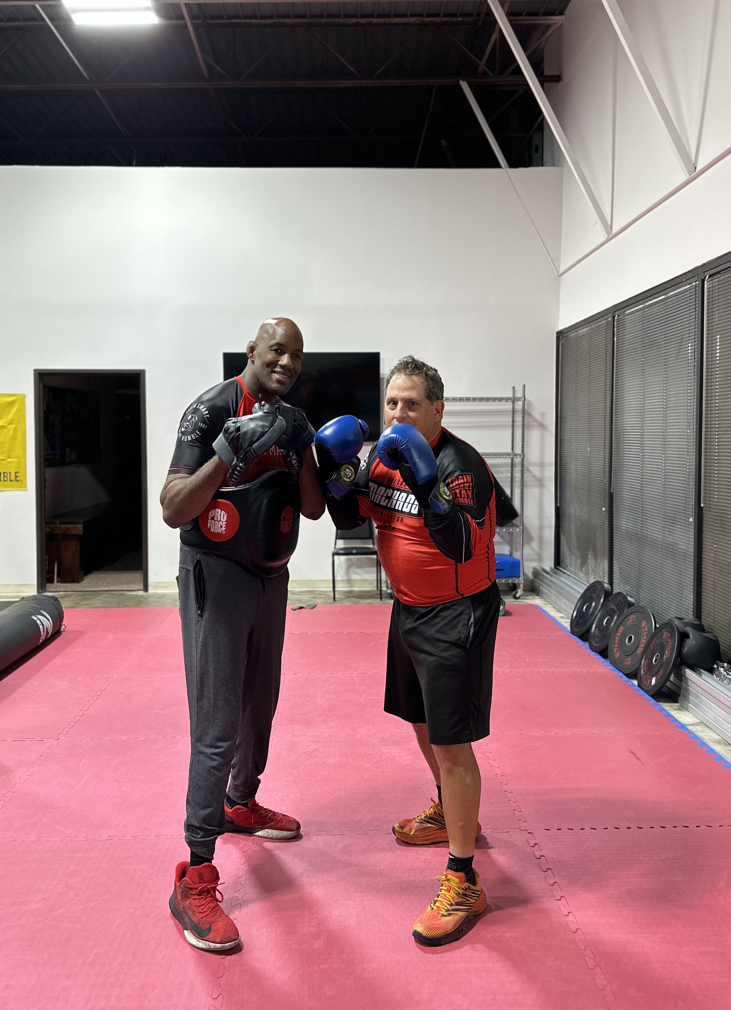 Two men in boxing gear and gloves posing inside a gym with pink mats, weights, and gym equipment in the background.
