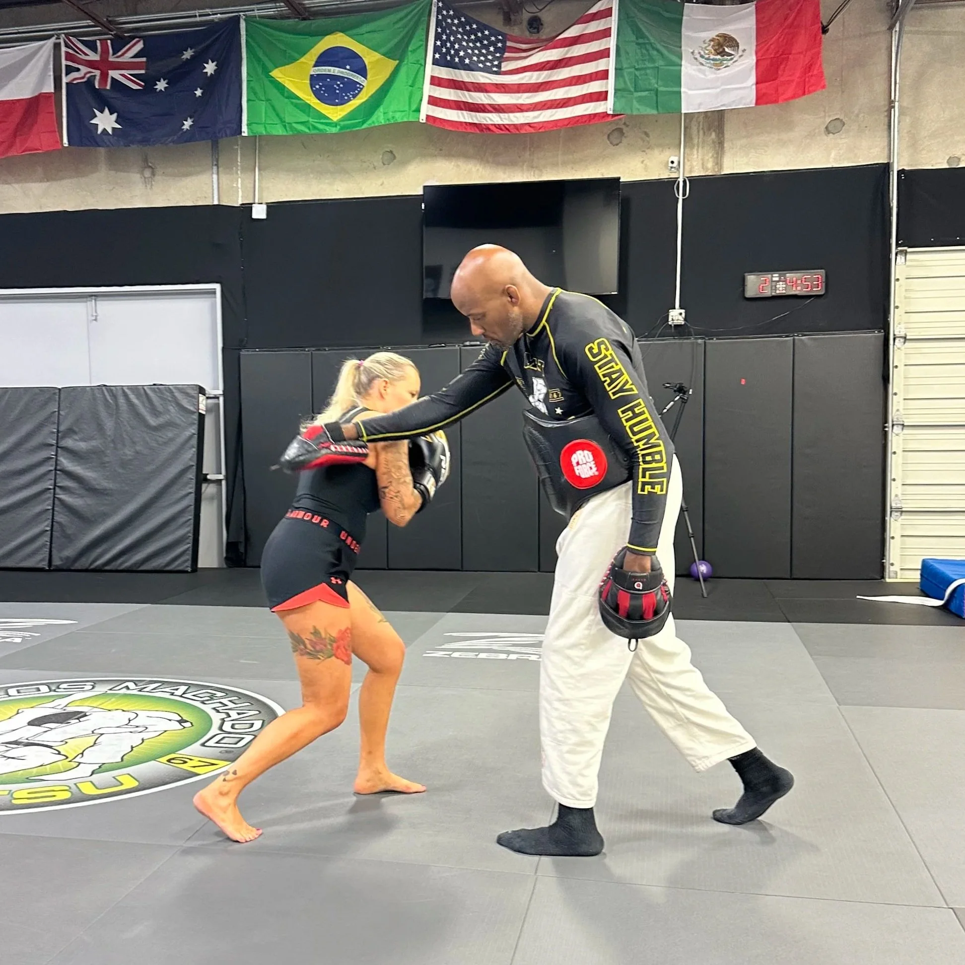 A woman practicing MMA with a trainer inside a gym. The woman is in a fighting stance, and the trainer is holding focus mitts for her to punch. Flags of Australia, Brazil, the United States, and Mexico hang on the wall in the background.