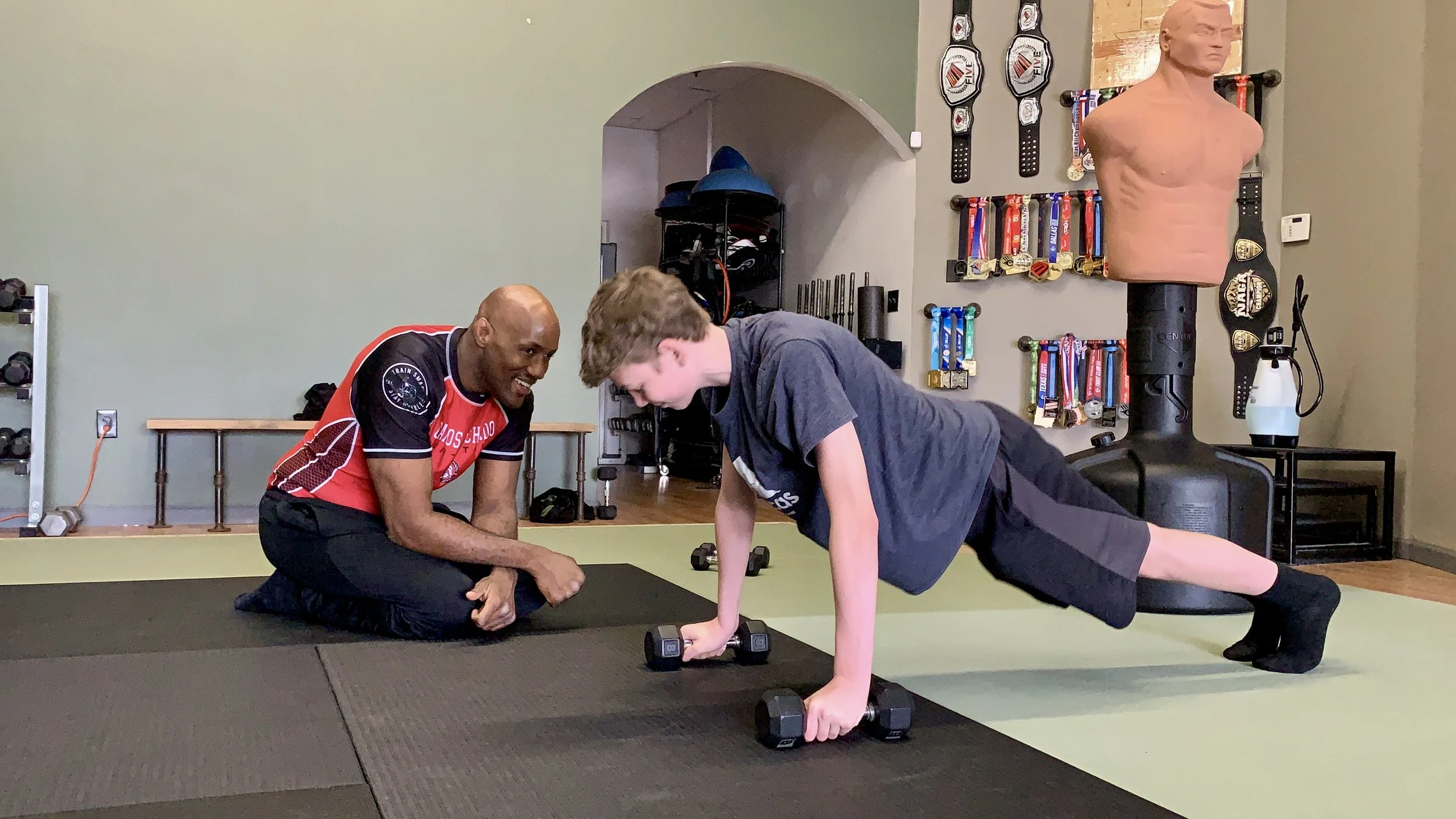 A young person performing a plank exercise using dumbbells while a male coach observes and encourages, in a gym with medals and boxing training equipment in the background.