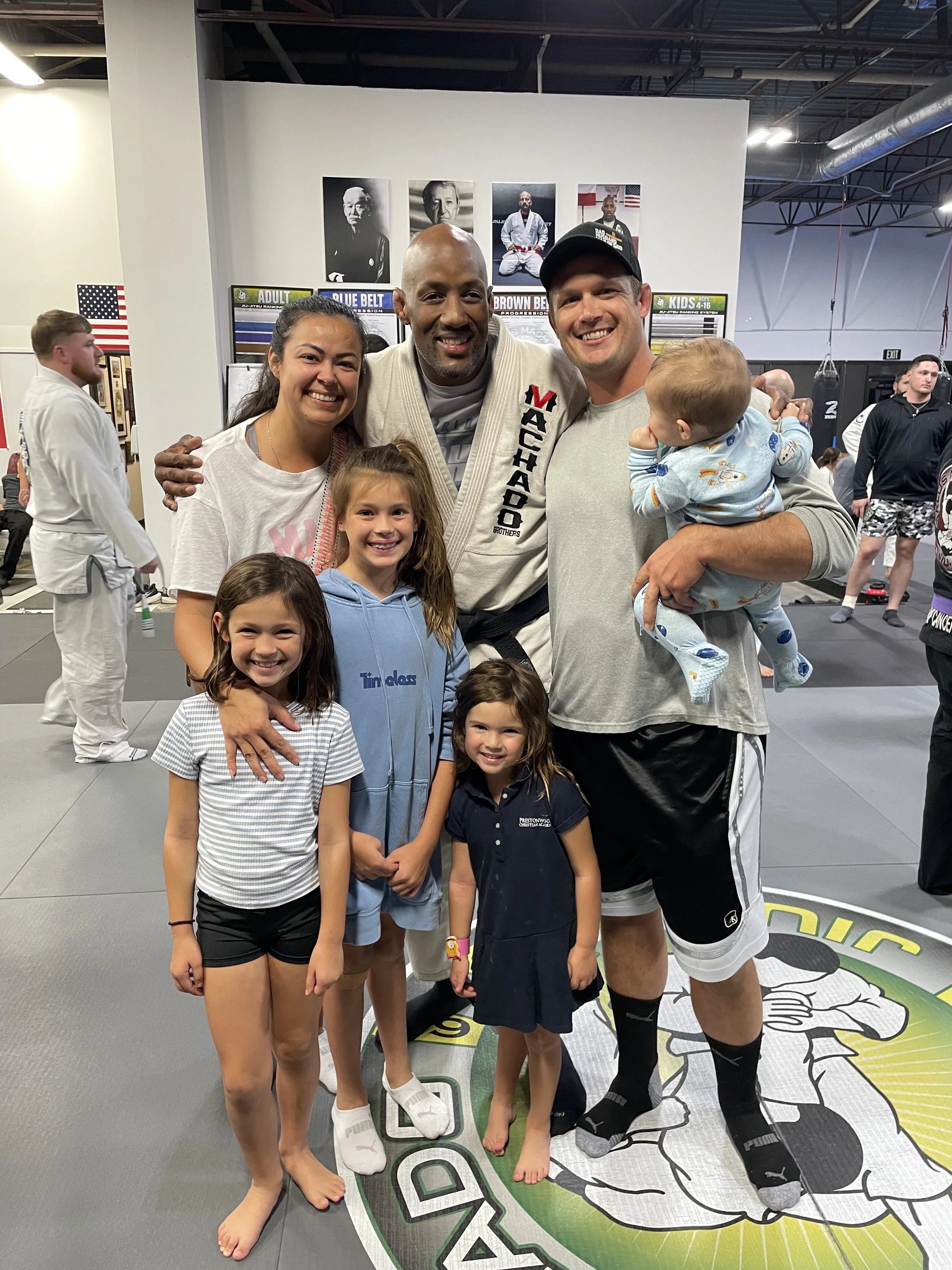 Group of seven people, including children and two adults, posing at a martial arts gym. One adult is wearing a gi with 'Vachado Brothers' on it, and the other is holding a small child. The background contains posters, flags, and several people practicing martial arts.