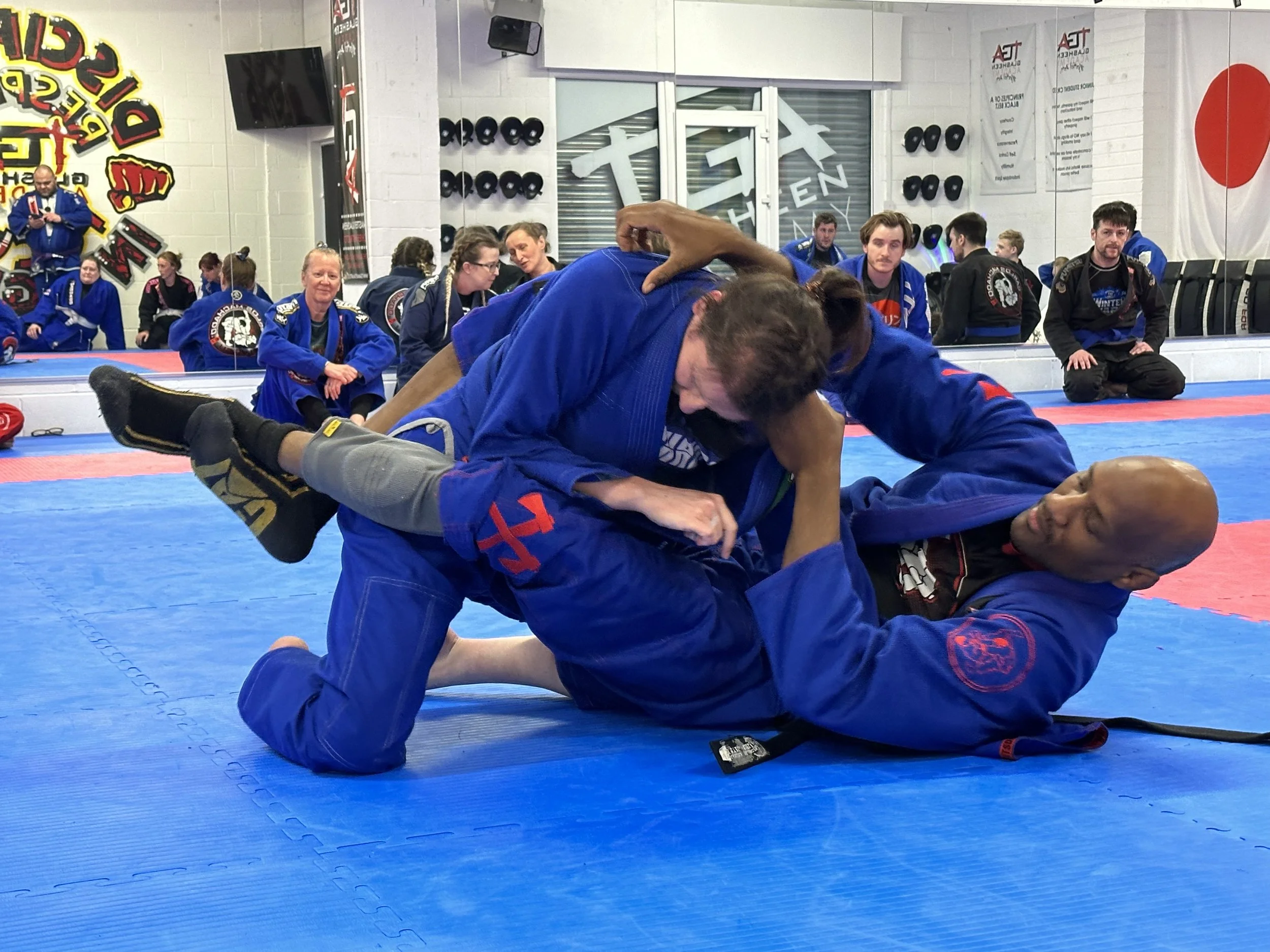 Two Brazilian Jiu-Jitsu practitioners in blue gis engaged in a grappling match on a blue mat, with people watching in the background.