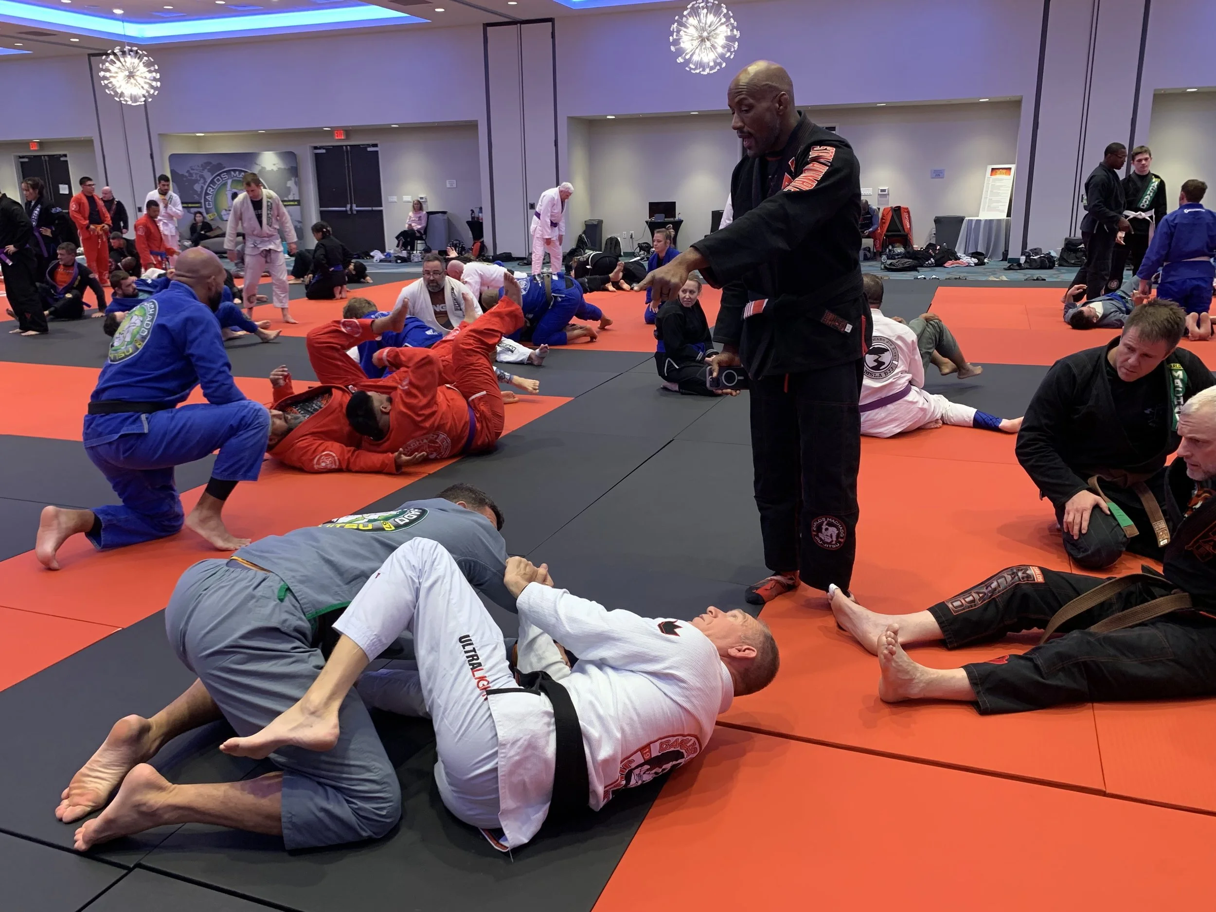 Brazilian Jiu-Jitsu practitioners in training inside a martial arts gym, with many people on orange and black mats practicing and receiving instruction.