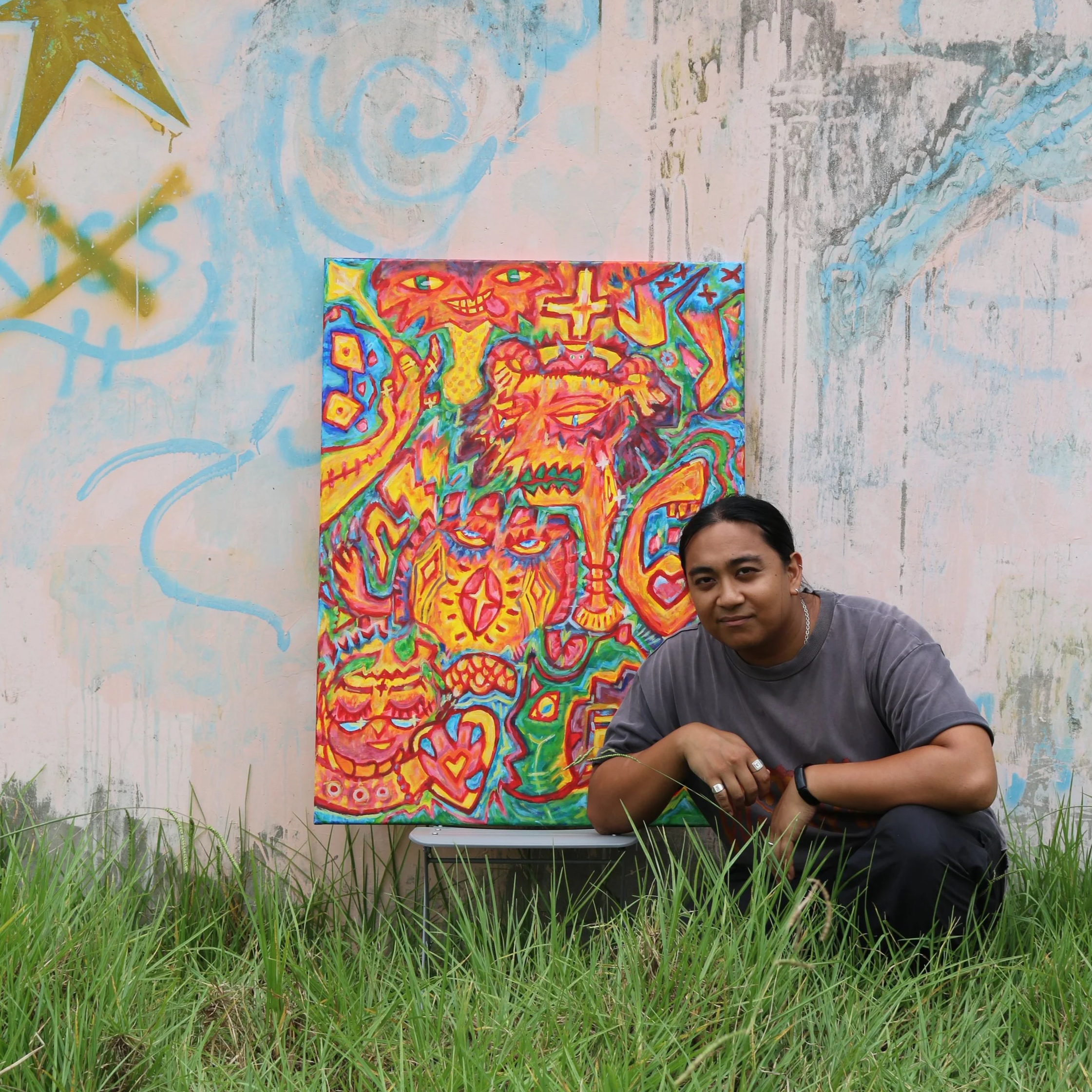 A man crouching in front of a colorful abstract painting outdoors, with grass in the foreground and a painted wall in the background.