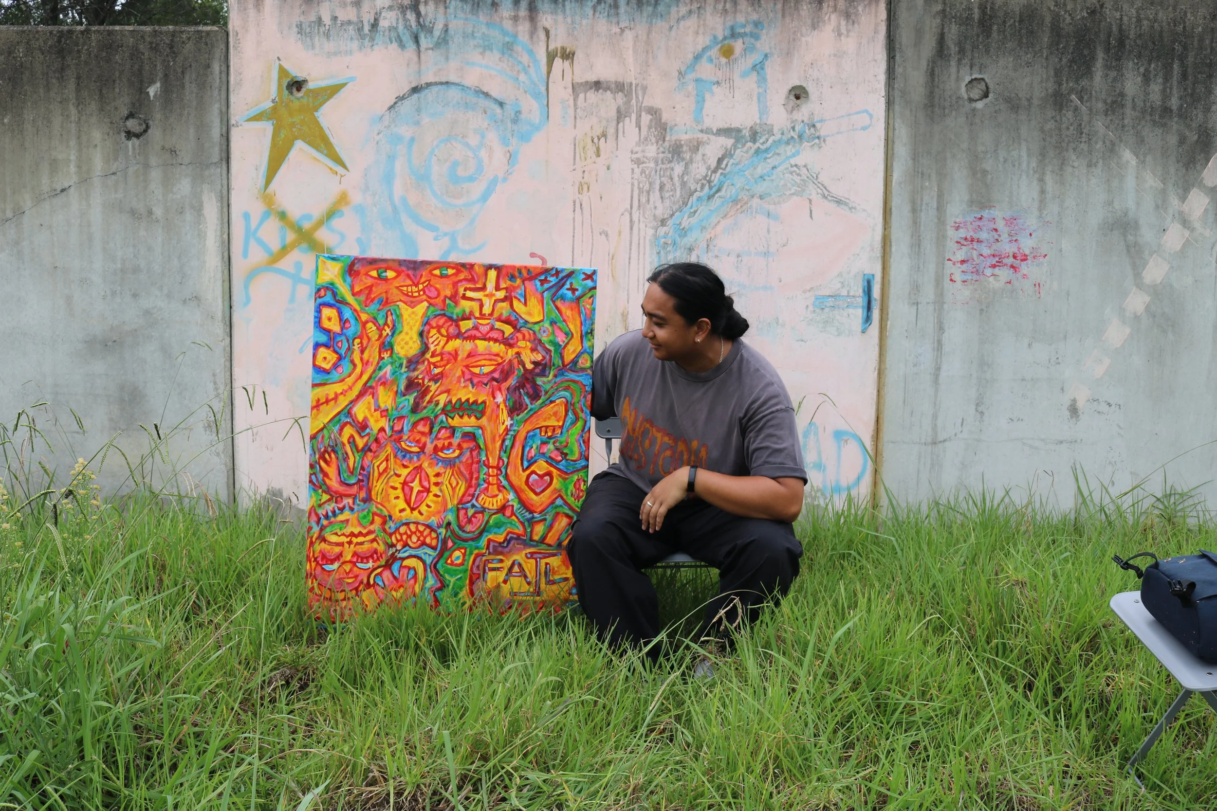 A person with dark hair wearing a gray T-shirt and black pants, sits in green grass in front of a colorful graffiti-style painting yellow, red, and orange colors. The person looks at the painting with a smile. Behind, there's a gray concrete wall with faded graffiti and a star decoration near the top left.