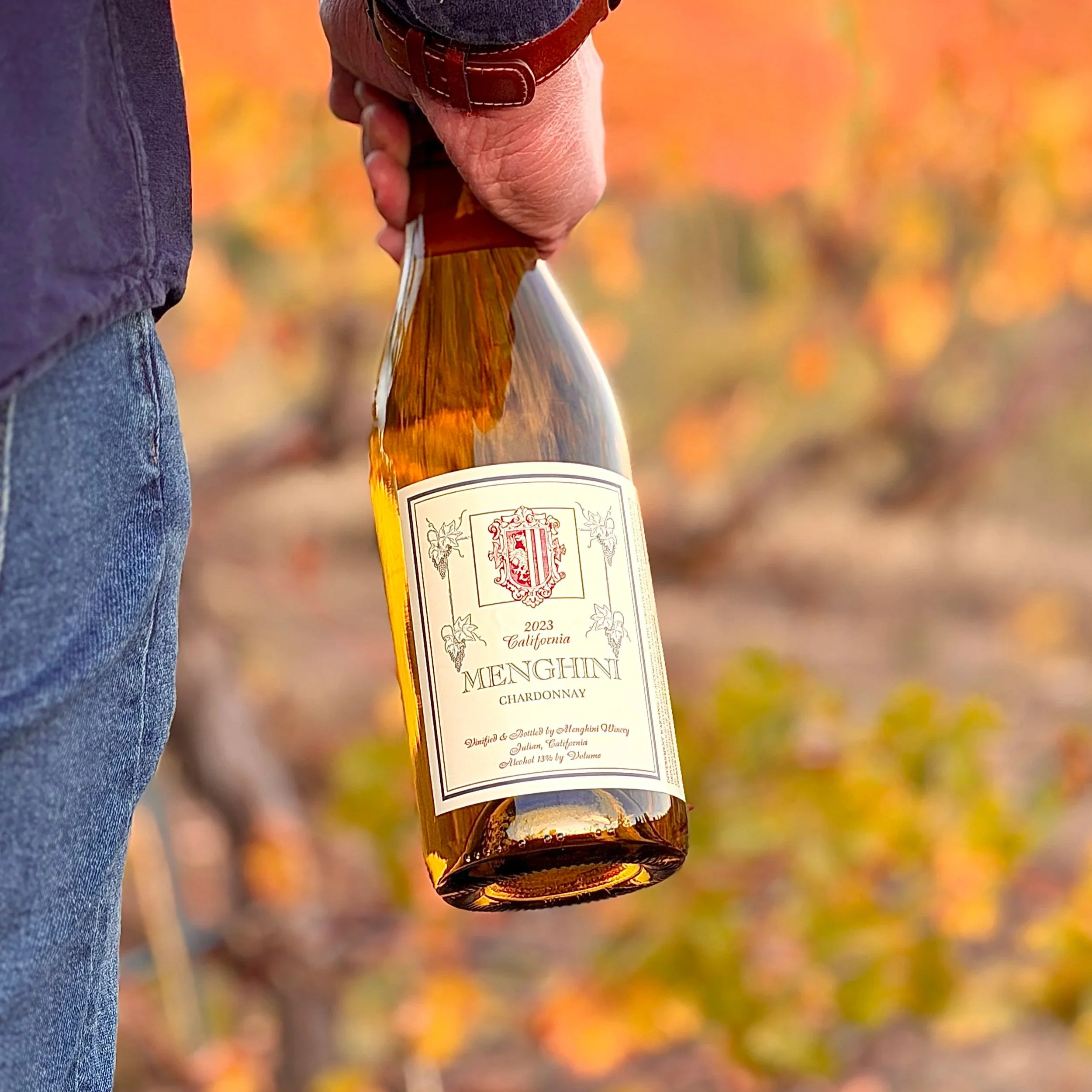 A bottle of Menghini Chardonnay wine outdoors with blurred autumn-colored foliage in the background.