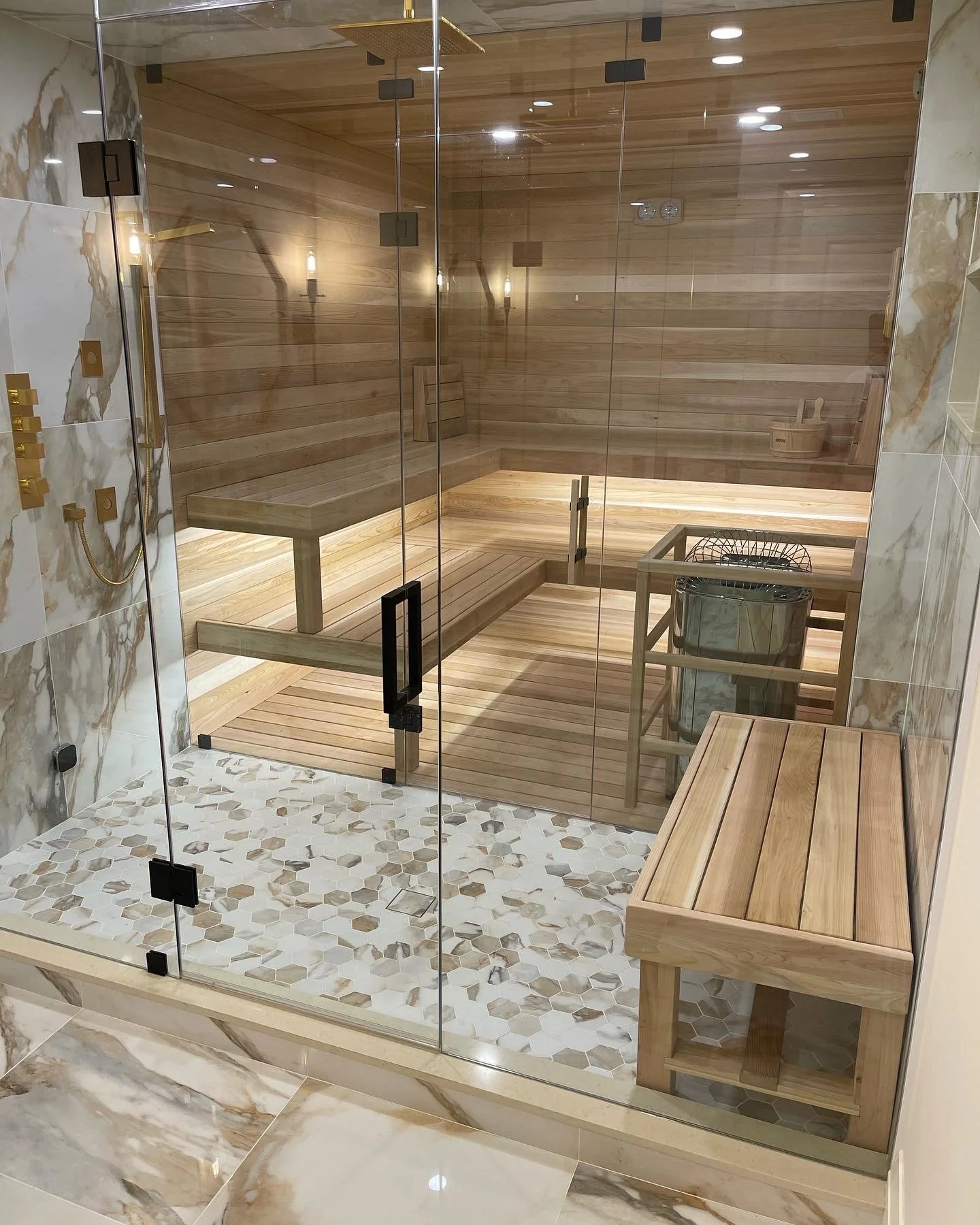 A glass-enclosed sauna with wooden interior panels, benches, a bucket, and a heater, with marble tiles on the outside wall and floor.'