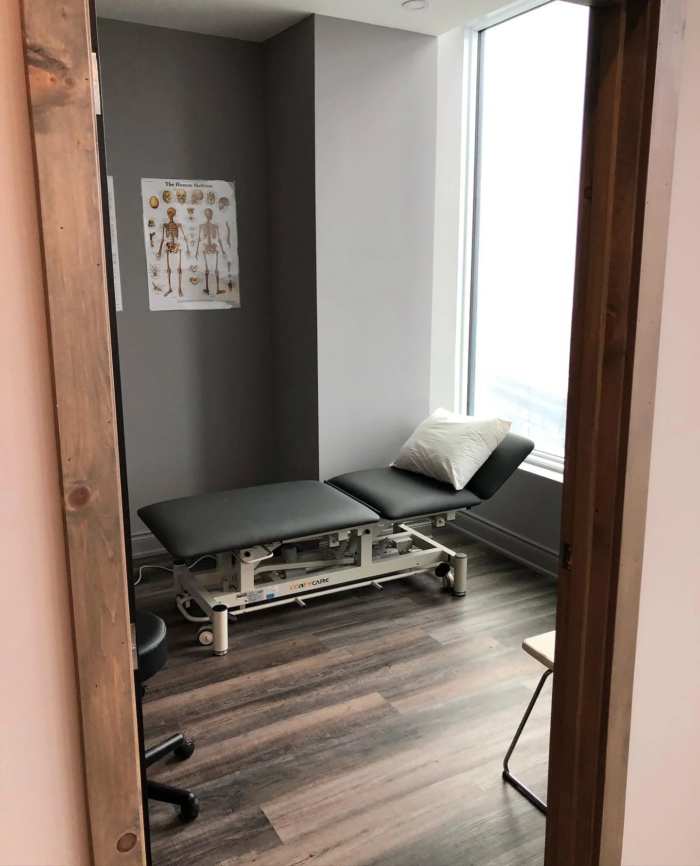 Completed renovation of a medical practitioner's room with an adjustable bed, a pillow, a window, and a poster of a human skeleton on the wall.