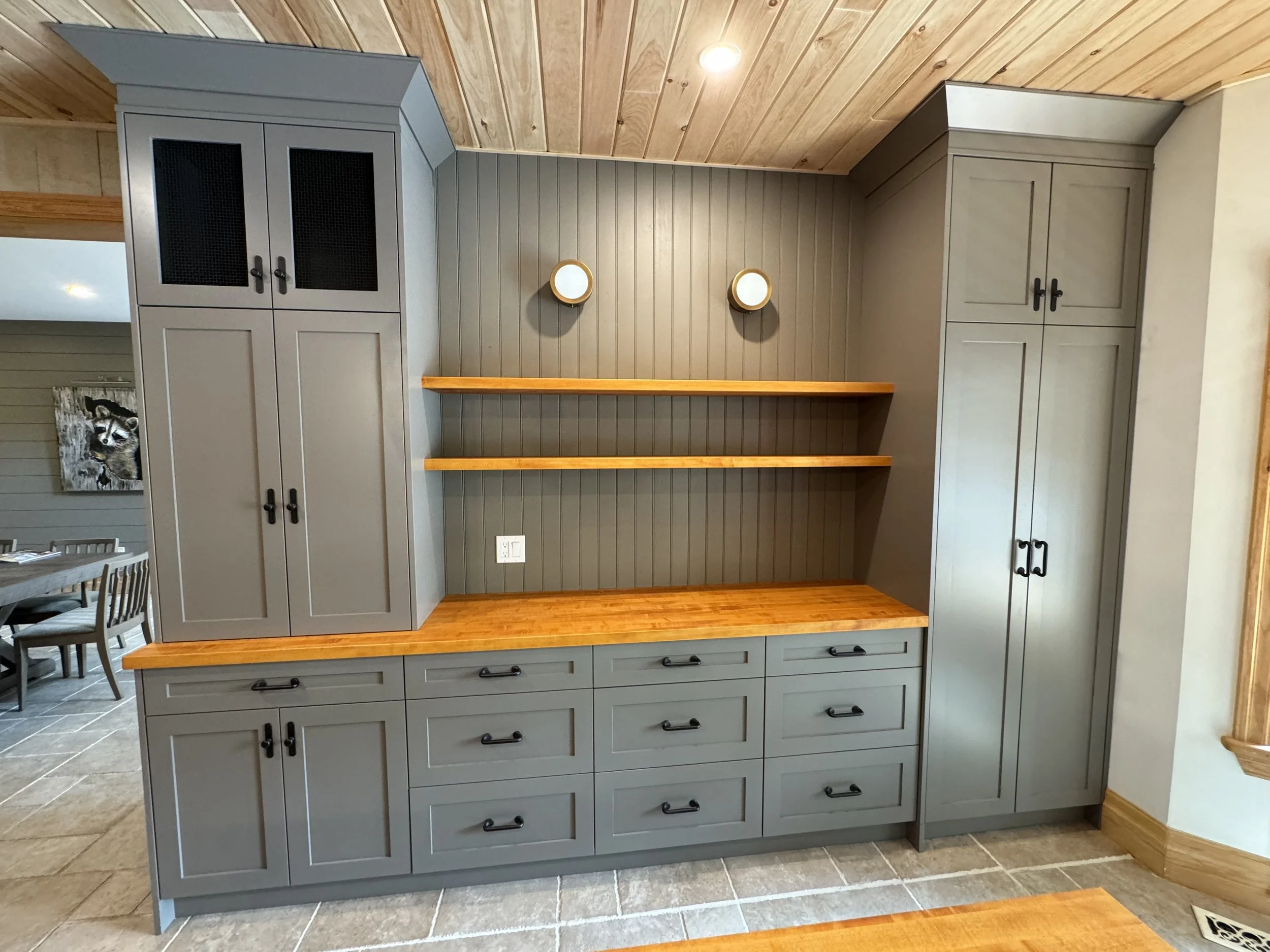 A gray built-in storage cabinet with black handles, featuring cabinets, drawers, and open wooden shelves in a room with wood-paneled walls and tile flooring.