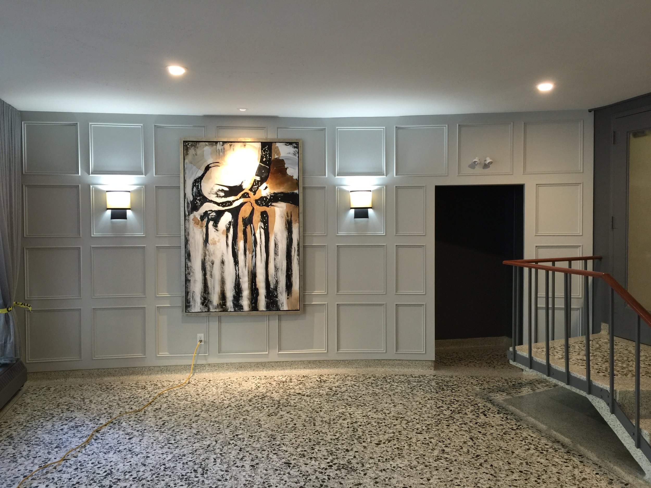 Interior view of a room with white paneled walls, abstract black and brown artwork, wall sconces with lights, a staircase with a wooden handrail, and a floor with speckled terrazzo tiles.