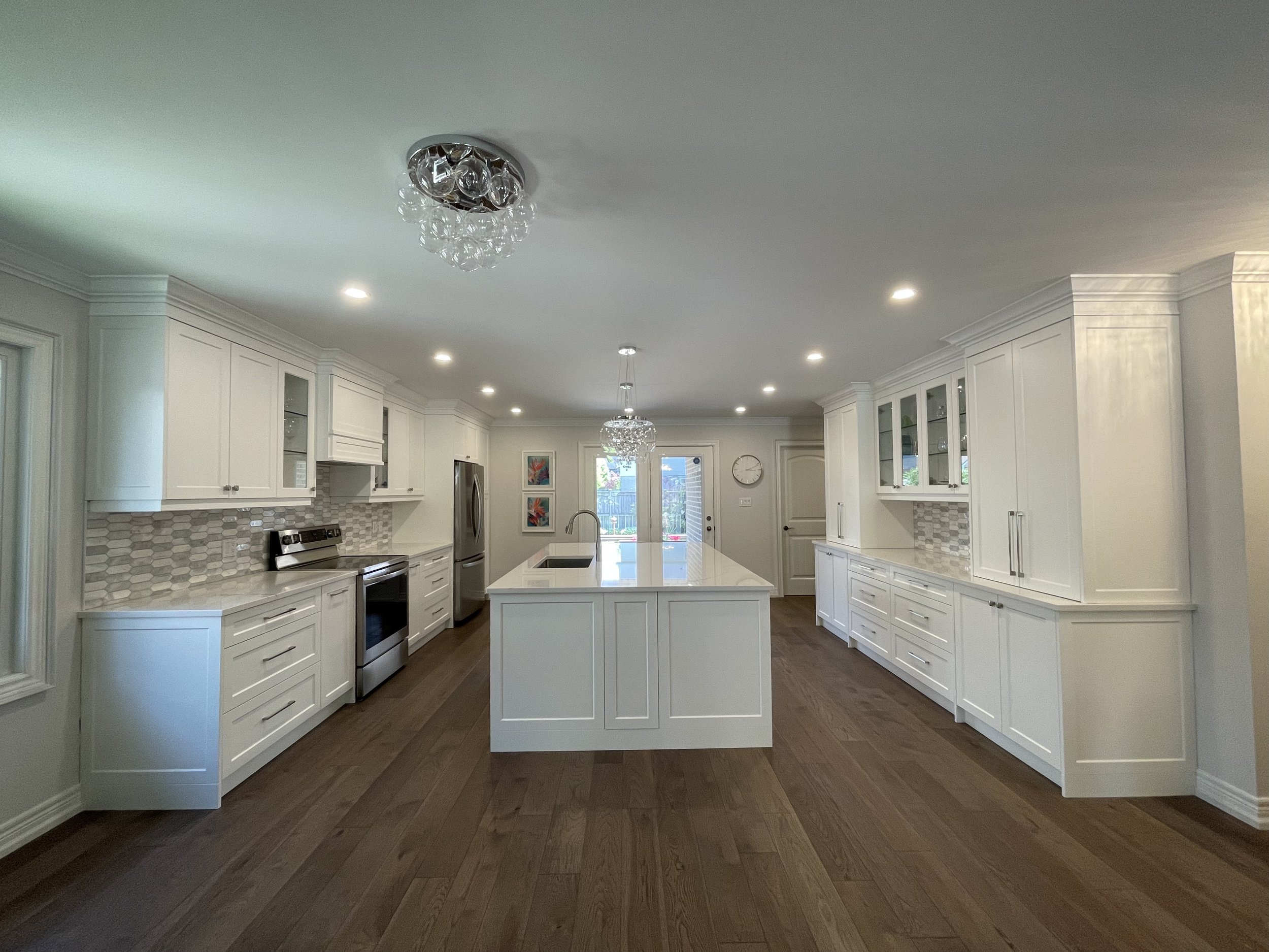 Modern white kitchen with hardwood floors, central island, stainless steel appliances, glass-front upper cabinets, and decorative lighting fixtures.