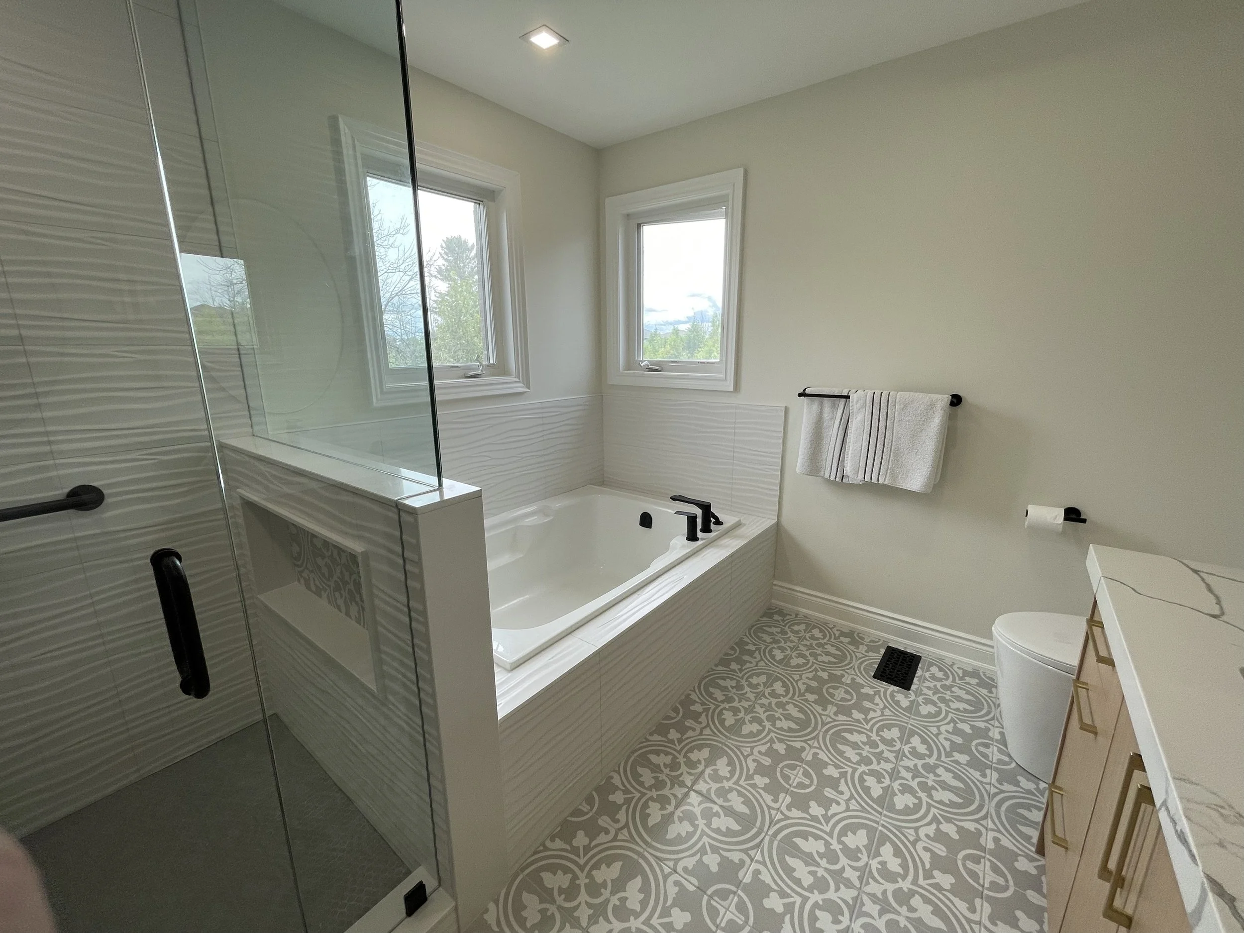 Modern bathroom with walk-in shower, bathtub, two windows, and decorative patterned floor tiles.