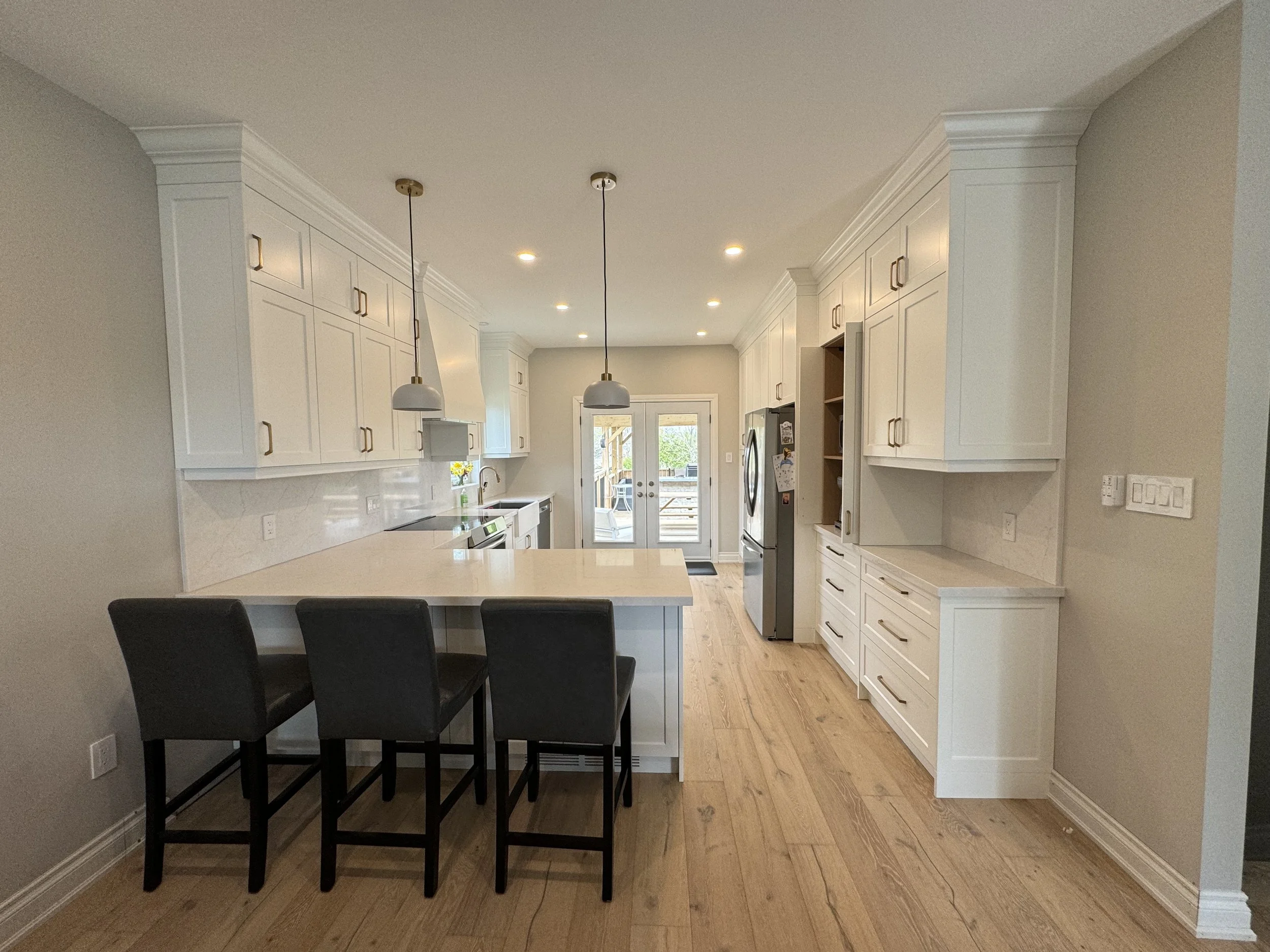 Modern white kitchen in Wasaga Beach, Ontario, with island, black chairs, stainless steel appliances, hardwood floors, and back door leading outside.