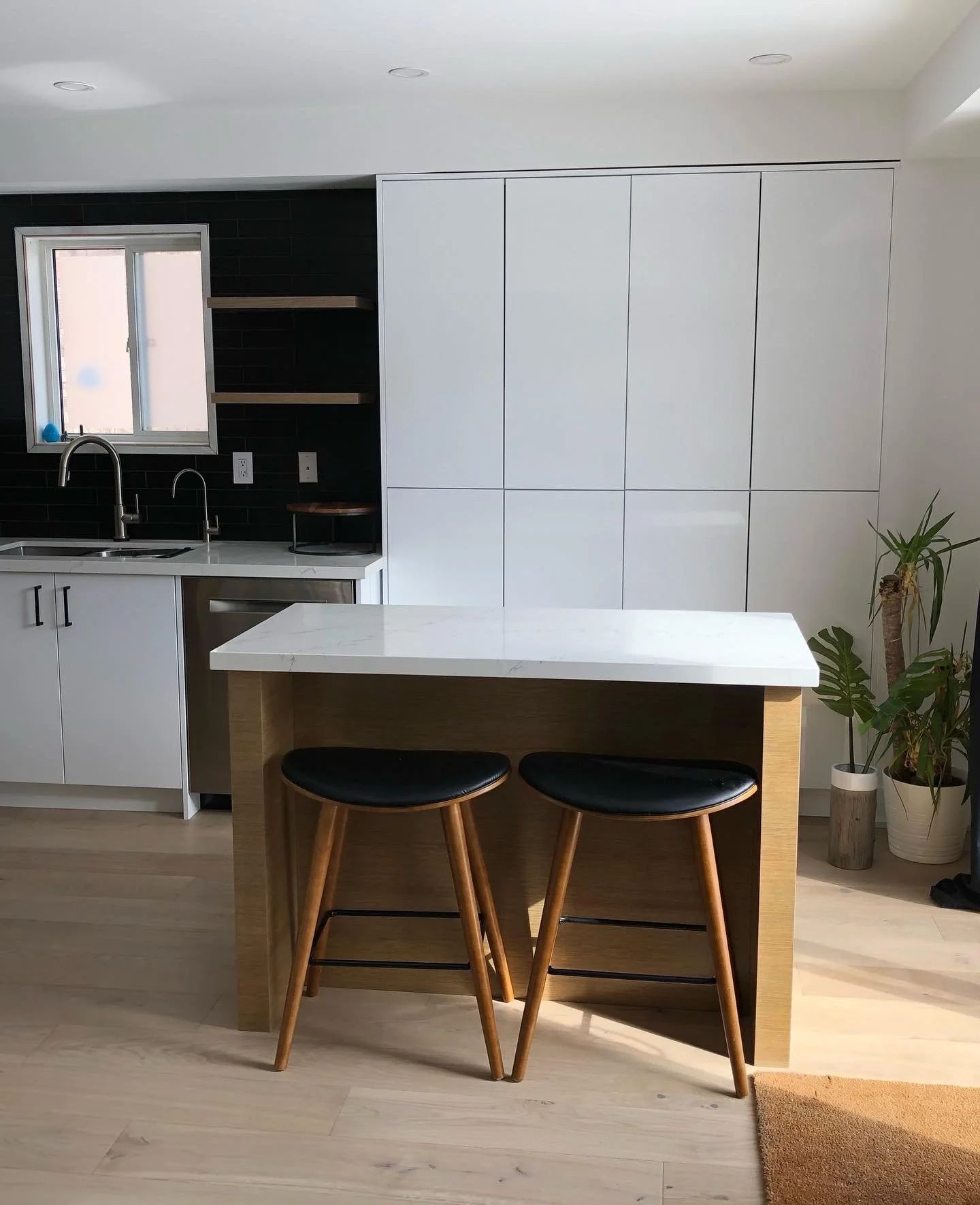 Modern kitchen with white cabinets, a small island with a white marble top, two black and wood bar stools, and a black tiled backsplash near a window.
