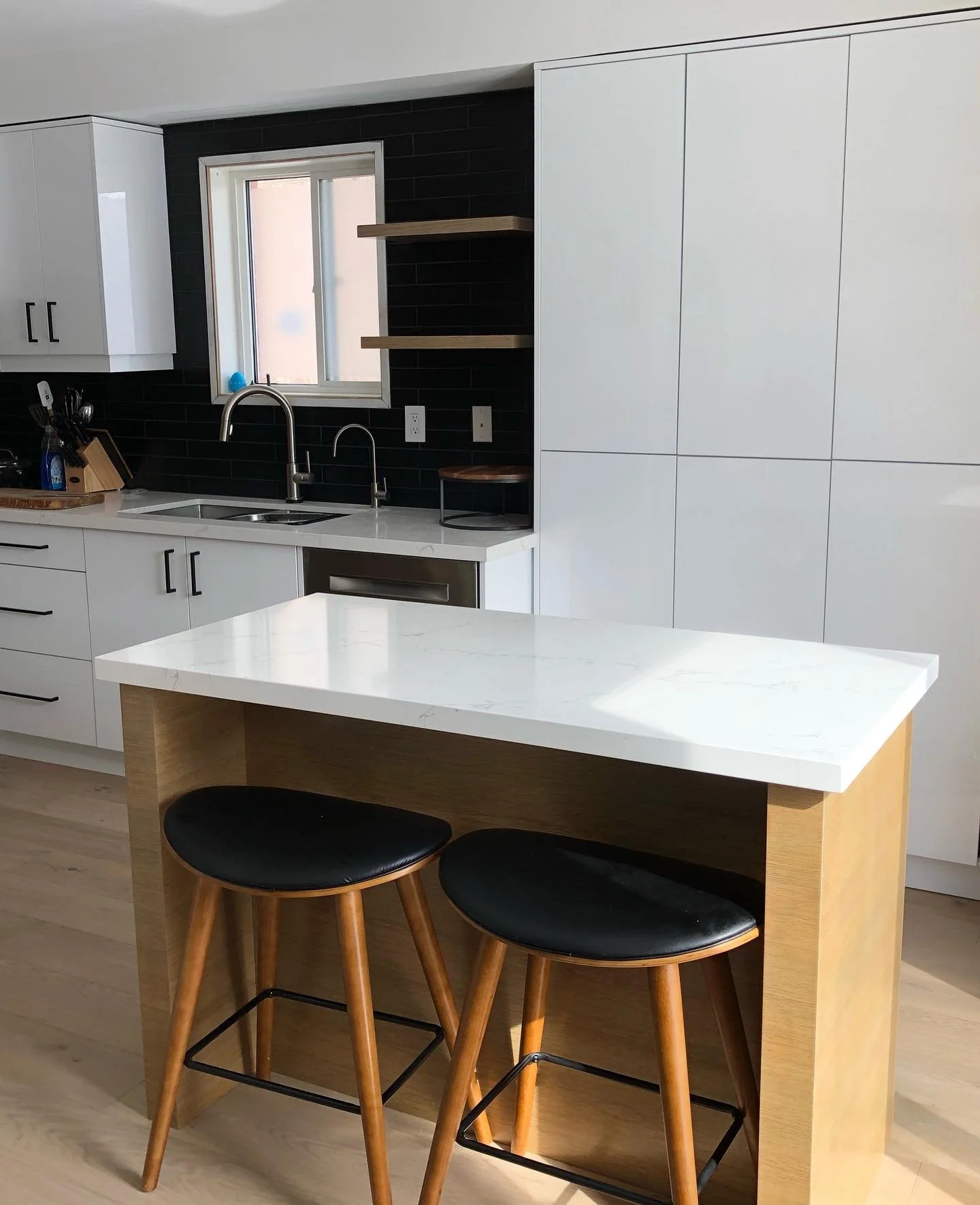 Modern kitchen with white cabinets, a black backsplash, a window over the sink, and an island with a white marble top and wooden base, accompanied by two black stool chairs.