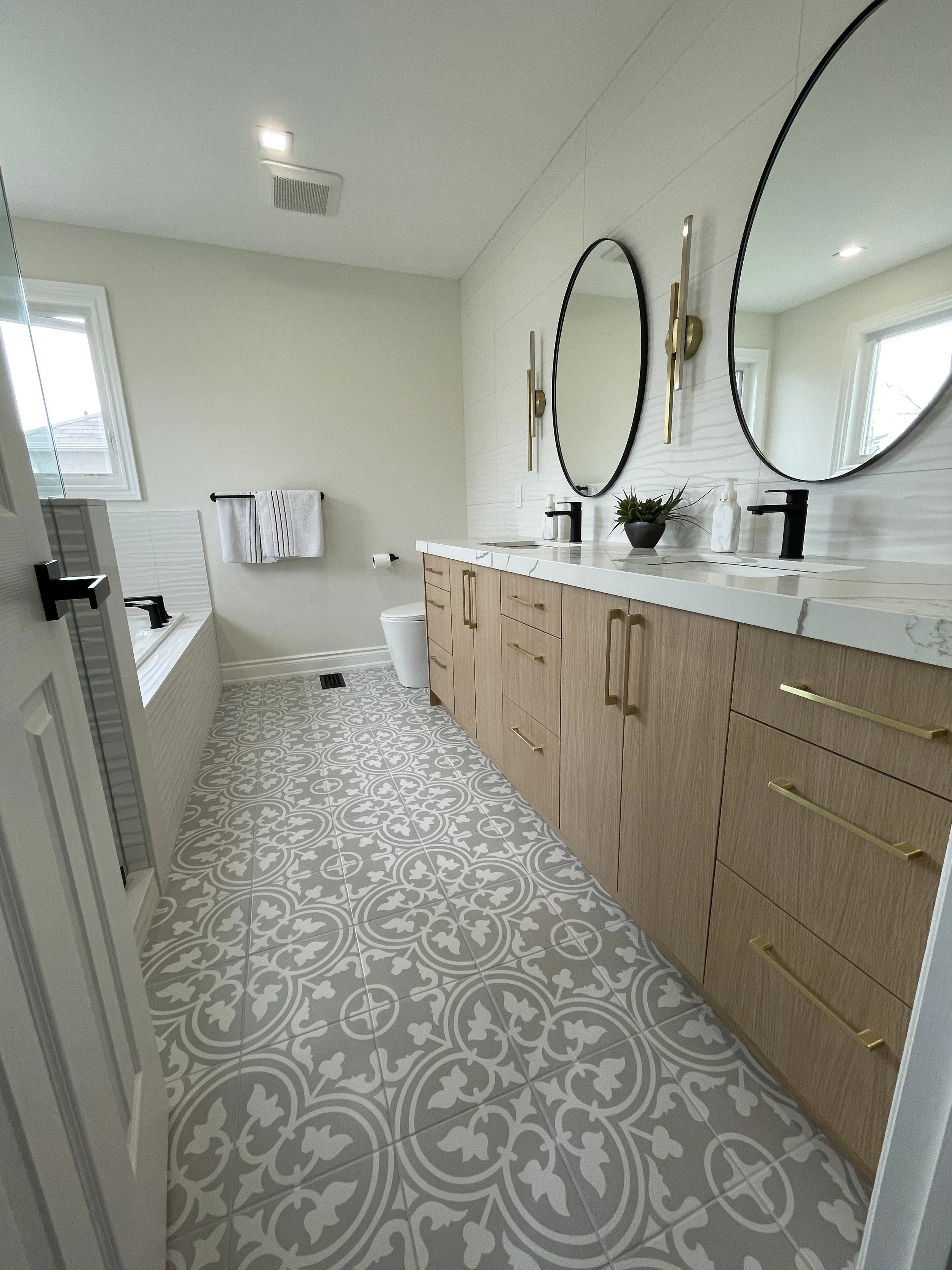 Modern bathroom with double vanity, large round mirrors, black faucets, beige wood cabinets, patterned gray and white floor tiles, a bathtub with a black faucet, a window letting in natural light, a towel rack with white towels, and decorative items 