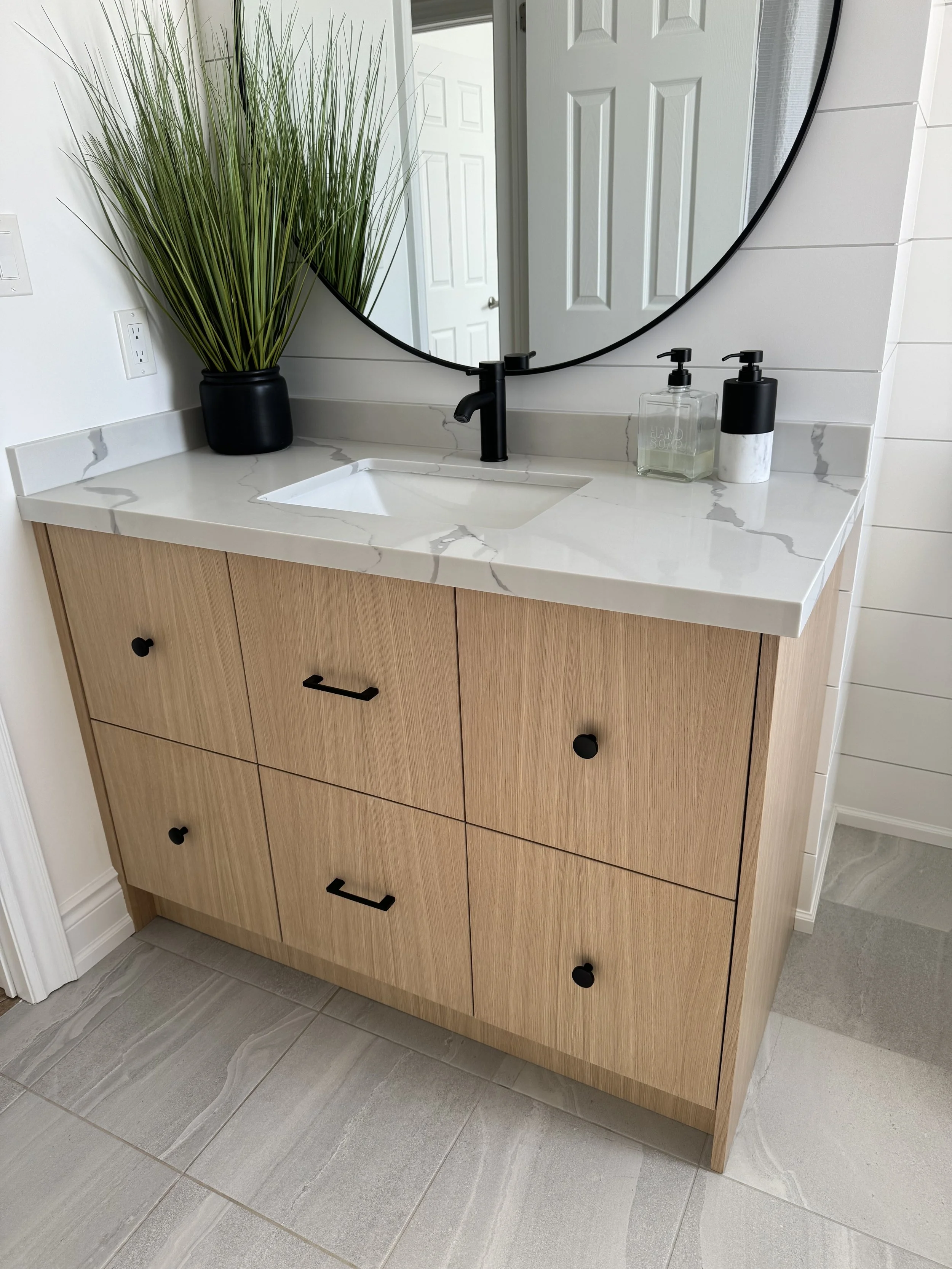 Bathroom vanity with a wooden cabinet, white marble countertop, black fixtures, large round mirror, green plant, hand soap, and lotion dispenser.