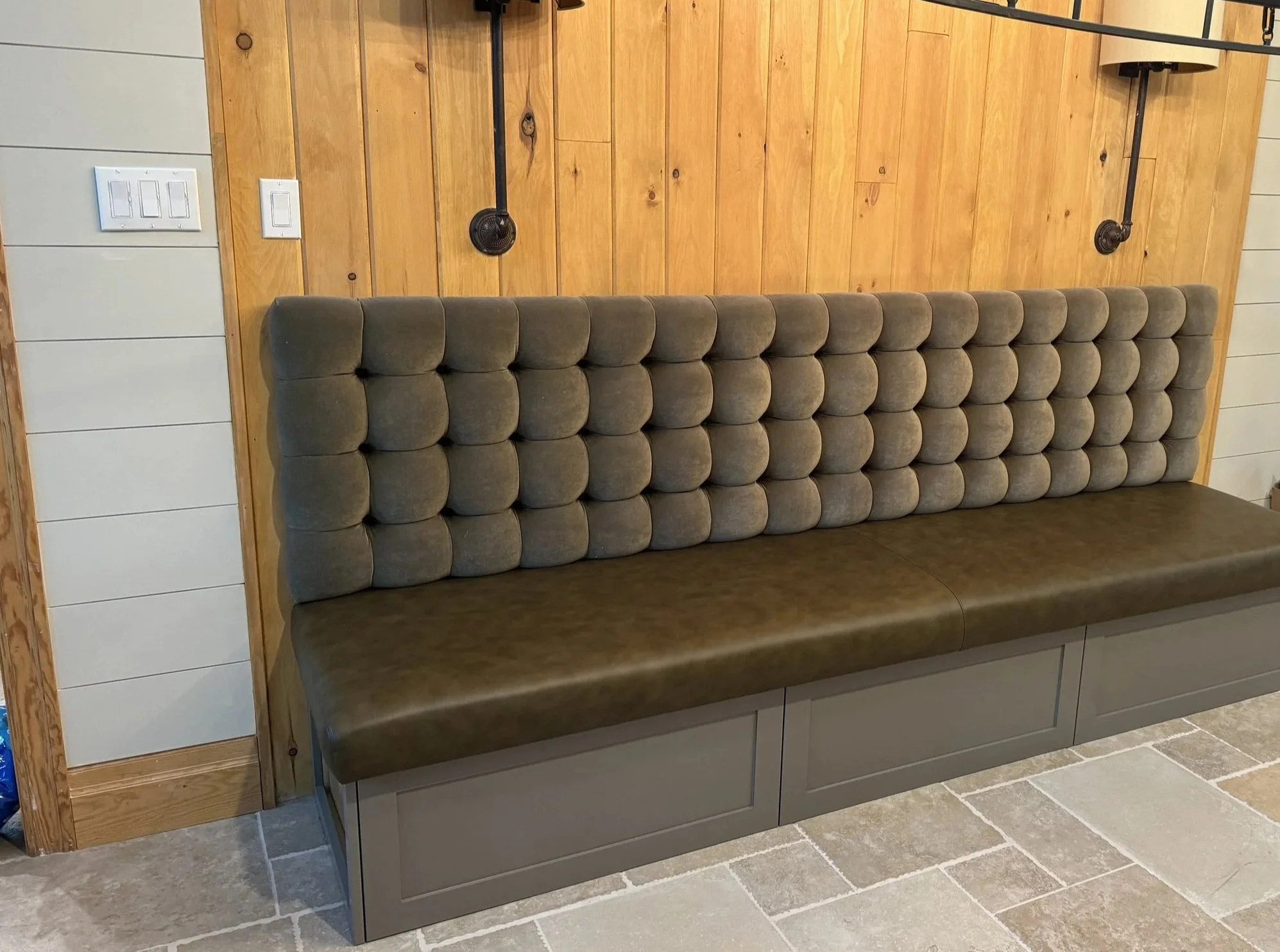 A long bench with tufted gray fabric backrest and a greenish cushion seat, placed against a wooden wall with two black wall-mounted lamps.