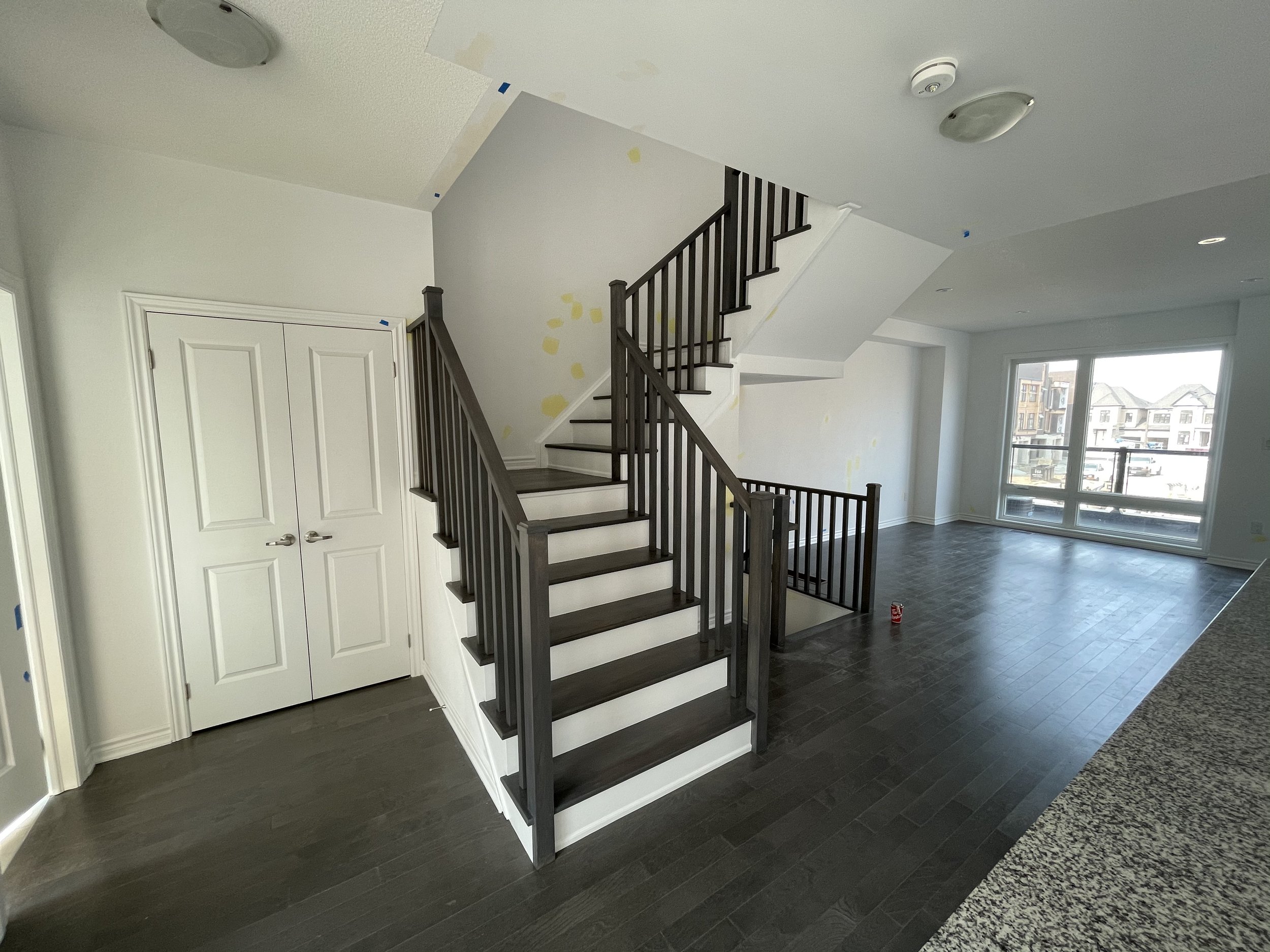 Empty living room with dark hardwood floors, a custom-built staircase with dark wood railing and white risers, large window with a view of neighboring buildings, and an open closet with double doors.