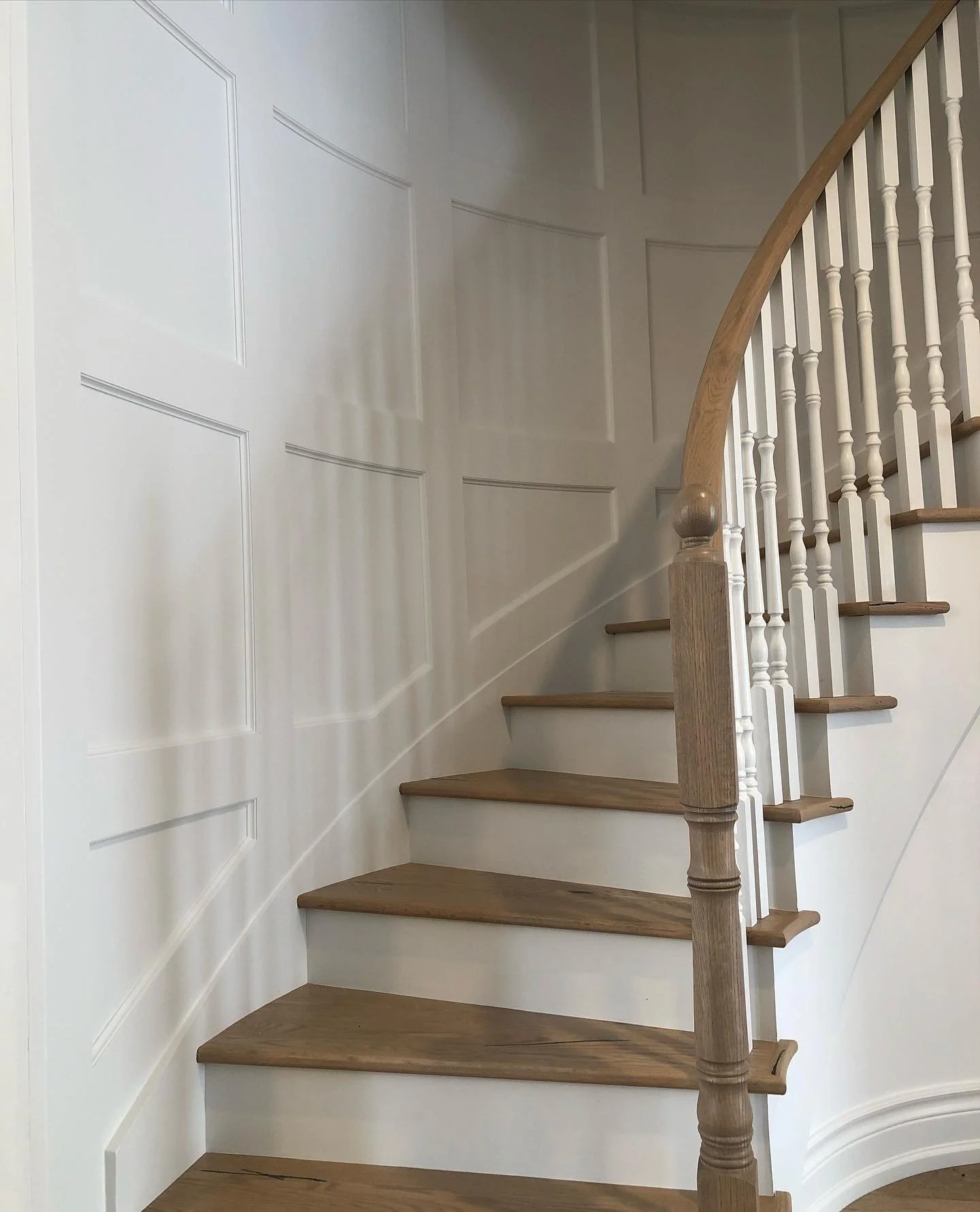 A curved staircase with wooden steps and white railings, against a white paneled wall.