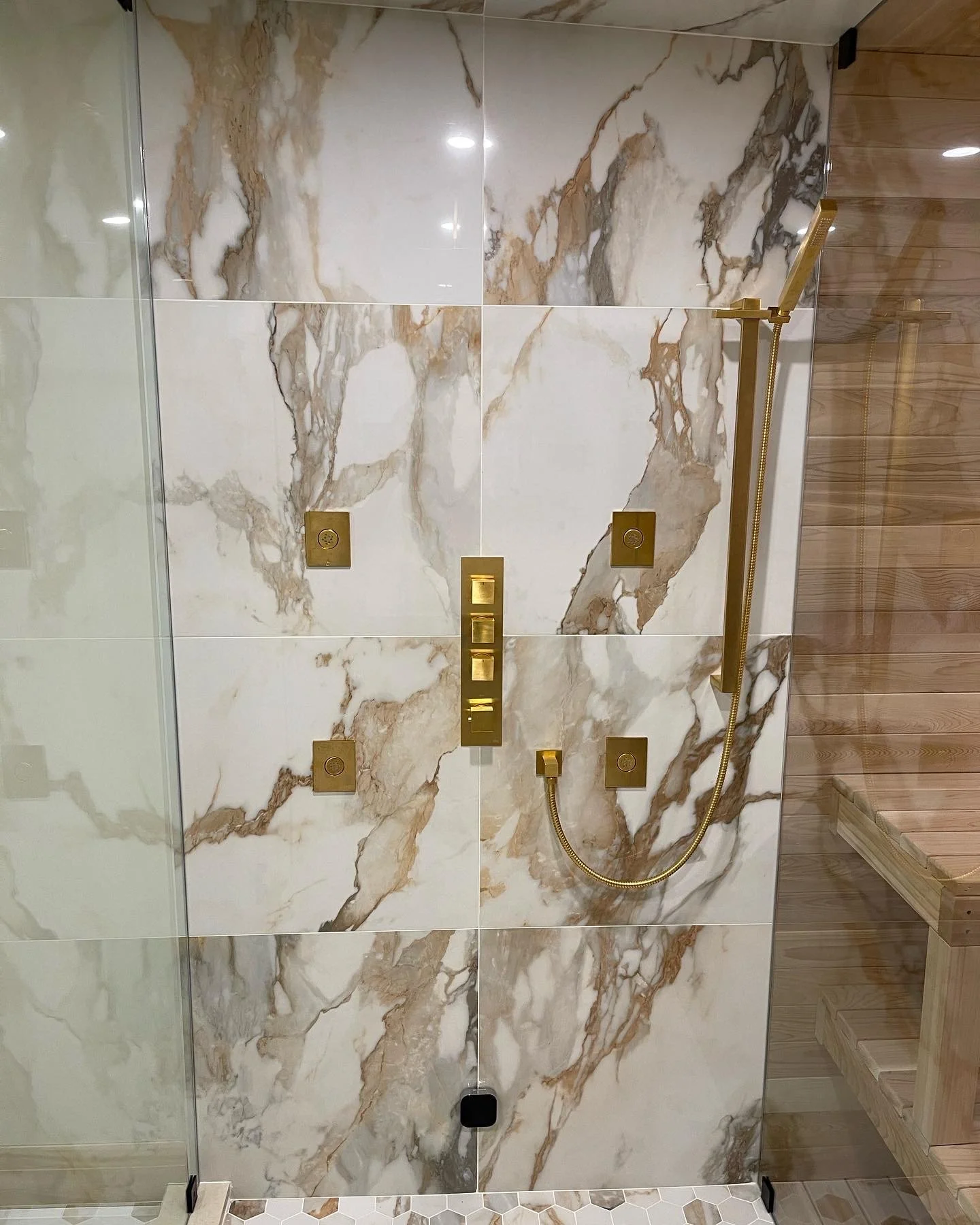 A marble shower wall with five gold-colored shower controls and a handheld showerhead with a flexible hose, adjacent to a wooden sauna bench.