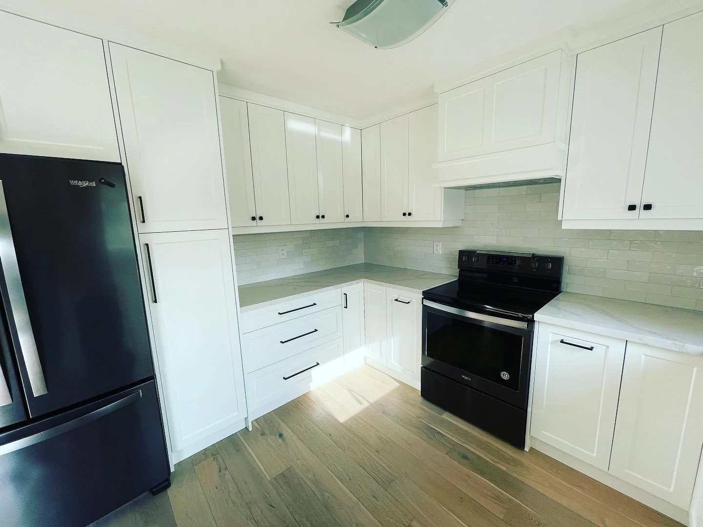 White kitchen with black appliances, including a refrigerator and an oven, white cabinets, and a beige subway tile backsplash, with light wood flooring.