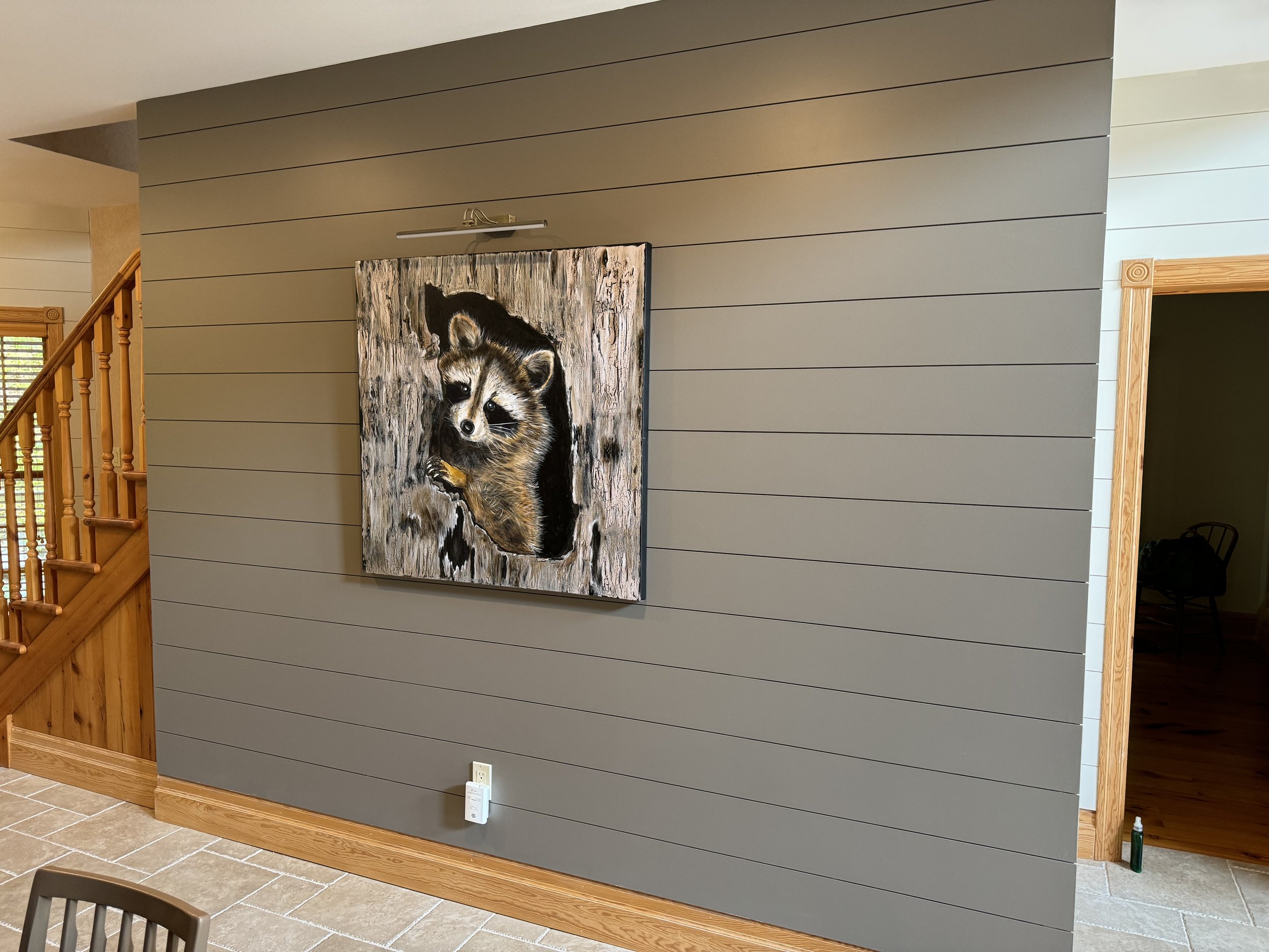 Painted artwork of a raccoon peeking out of a tree hollow on a gray wall in a room with wood trim.