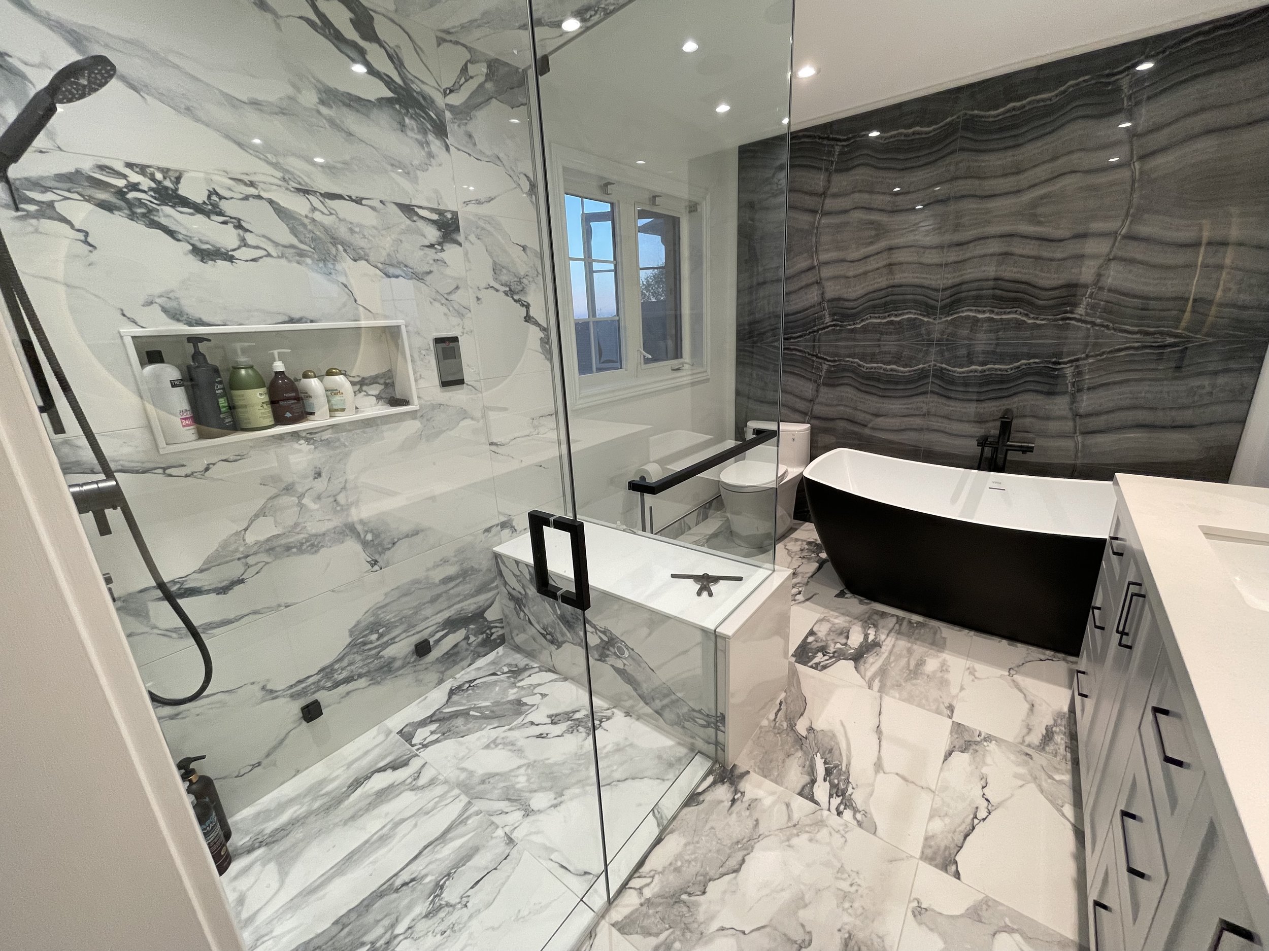 Modern Barrie bathroom with marble floors and walls, a walk-in shower with glass enclosure, a black and white bathtub, a white vanity with a sink, and a window with a view outside.