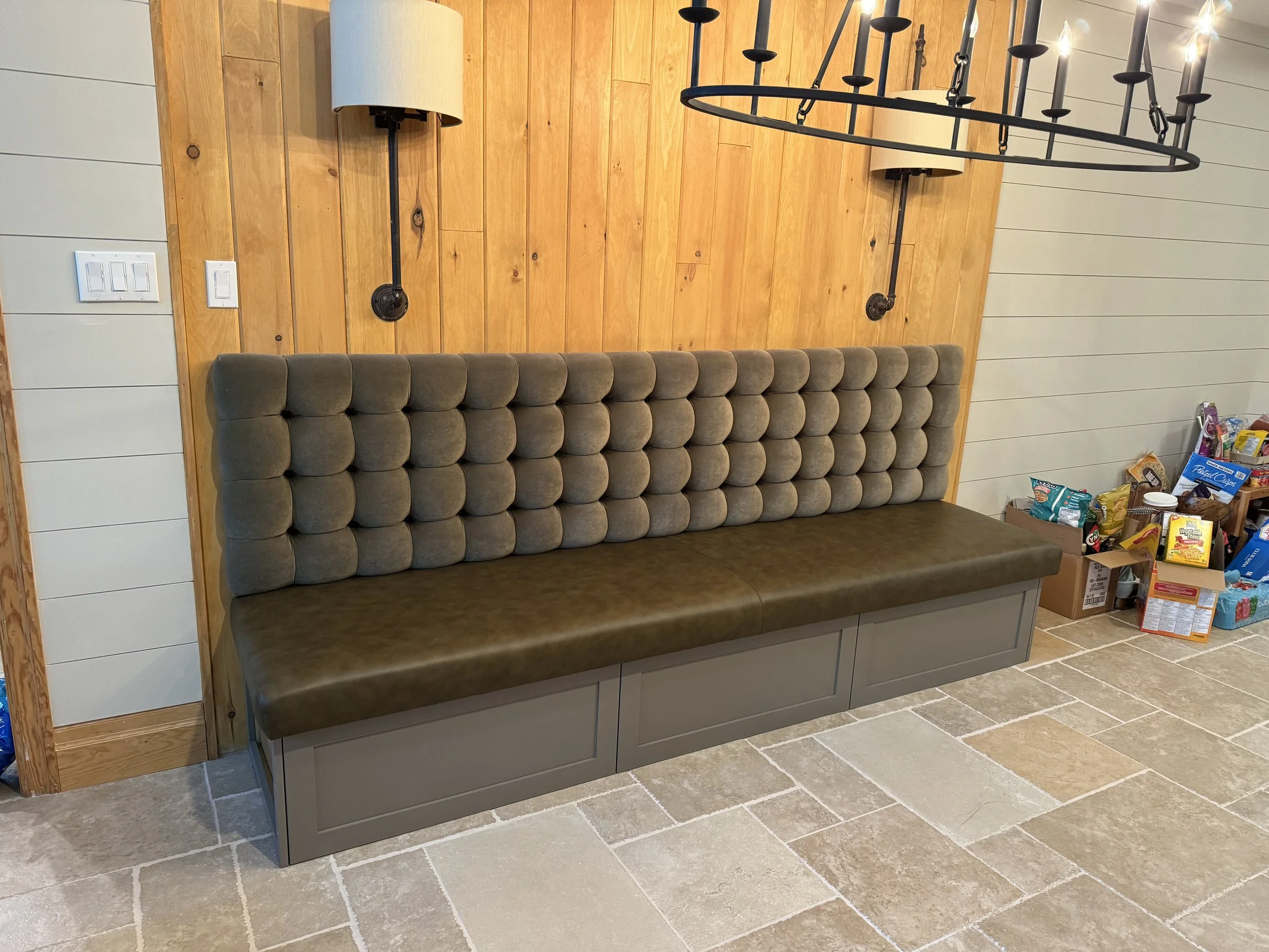 A built-in bench with a tufted backrest and storage drawers at the bottom, set against a wooden-paneled wall in a room with tiled flooring and boxes of snacks to the right.
