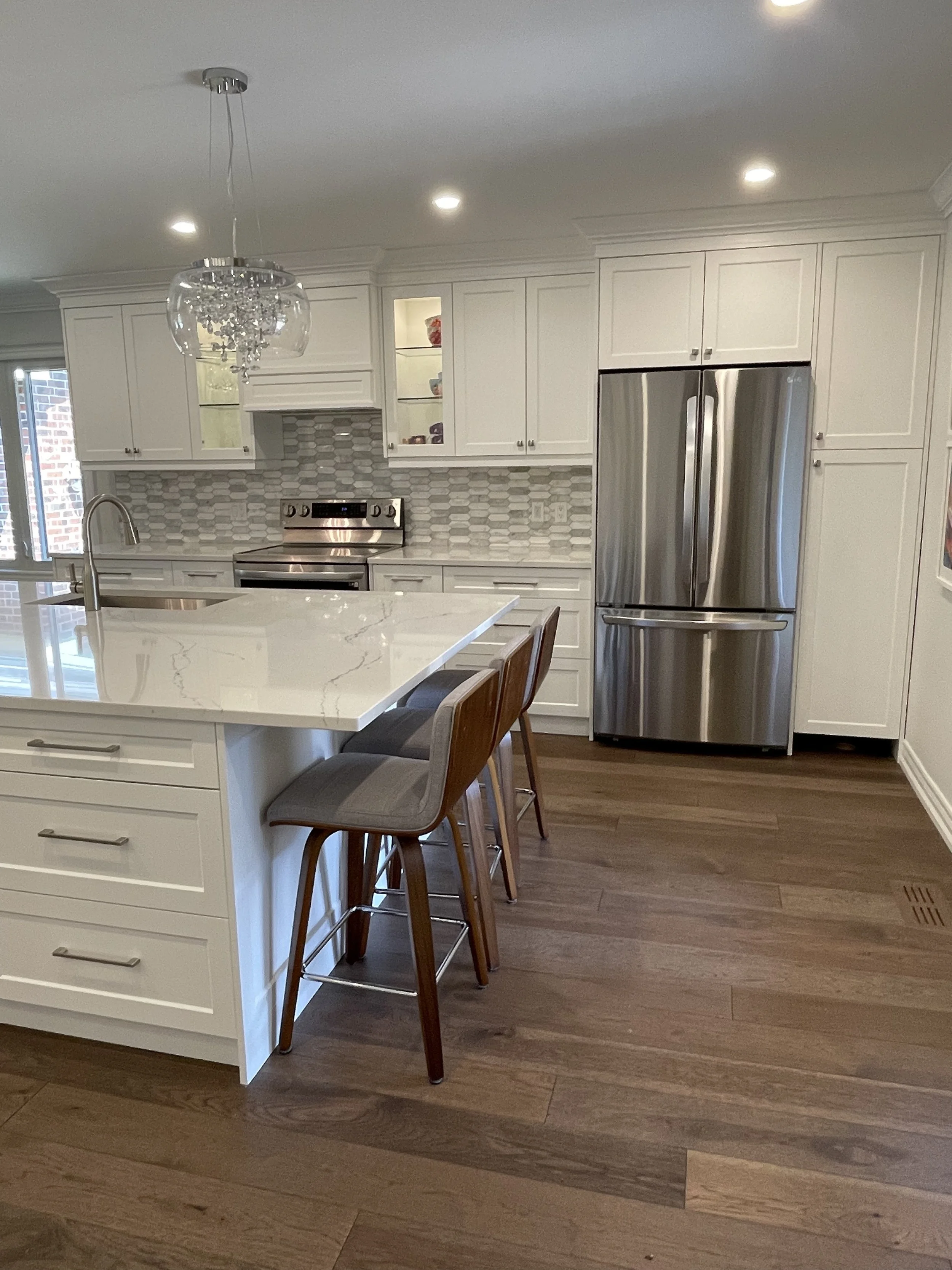 Modern kitchen with white cabinets, stainless steel refrigerator and stove, marble island with bar stools, wood flooring, and a decorative chandelier.