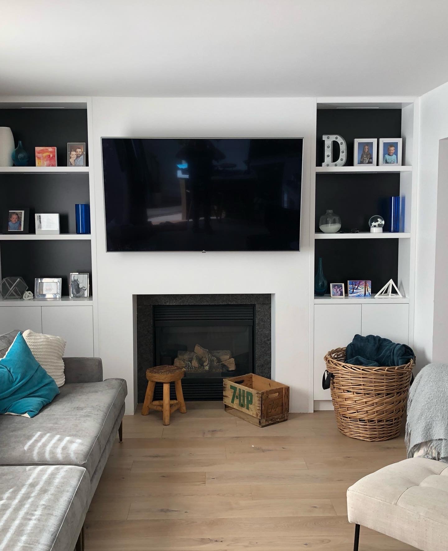 Living room custom renovation with a wall-mounted TV above a fireplace, surrounded by white shelves with family photos and decorative items, a small wooden stool, a box labeled 7-UP, a laundry basket, and couches with cushions.