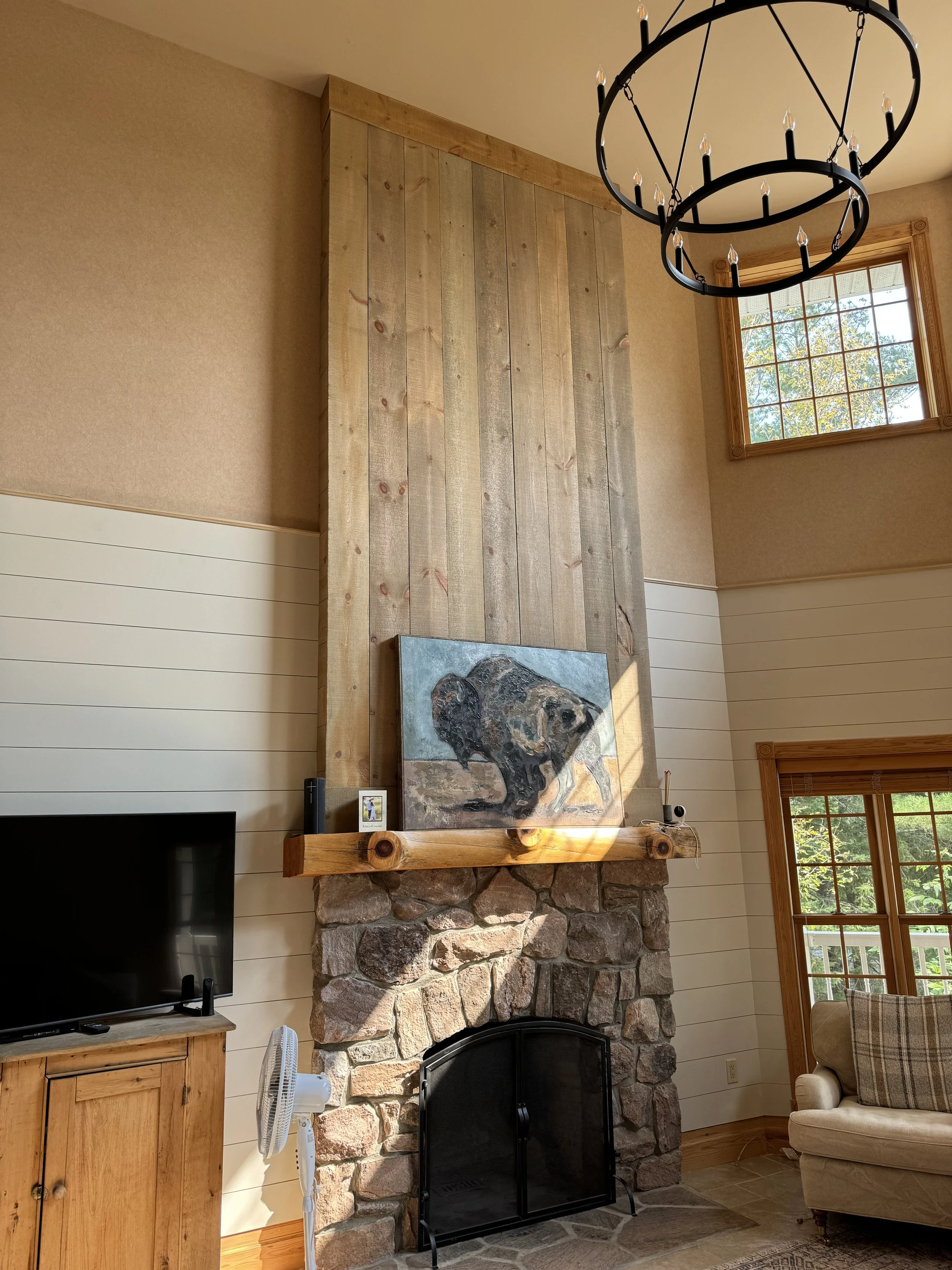 Living room with stone fireplace, wooden mantel, painting of a bear, television, fan, chandelier, and windows with wooden trim.