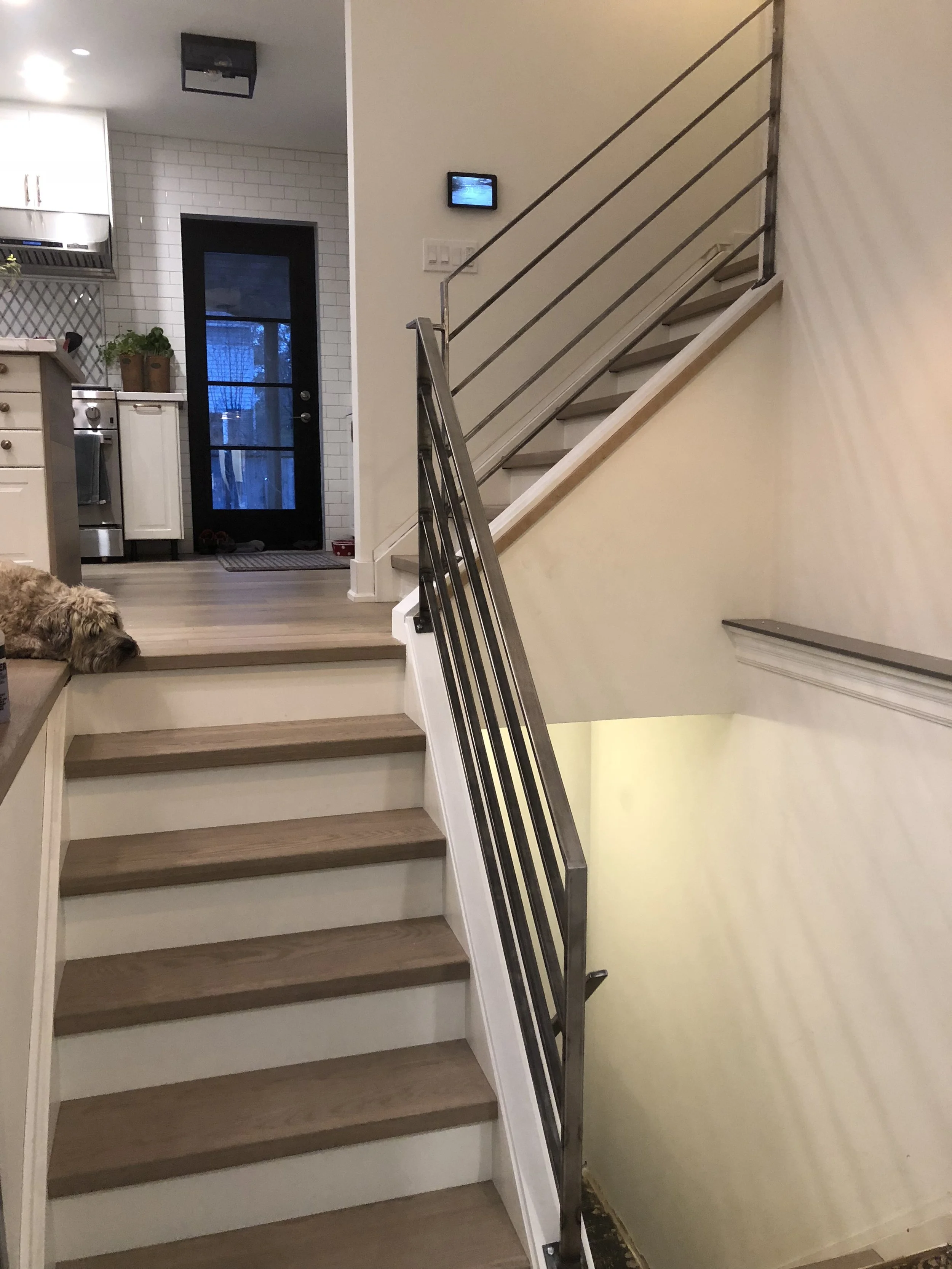 Living room with staircase, a dog lying on the stairs, kitchen in the background, and a black door leading outside.