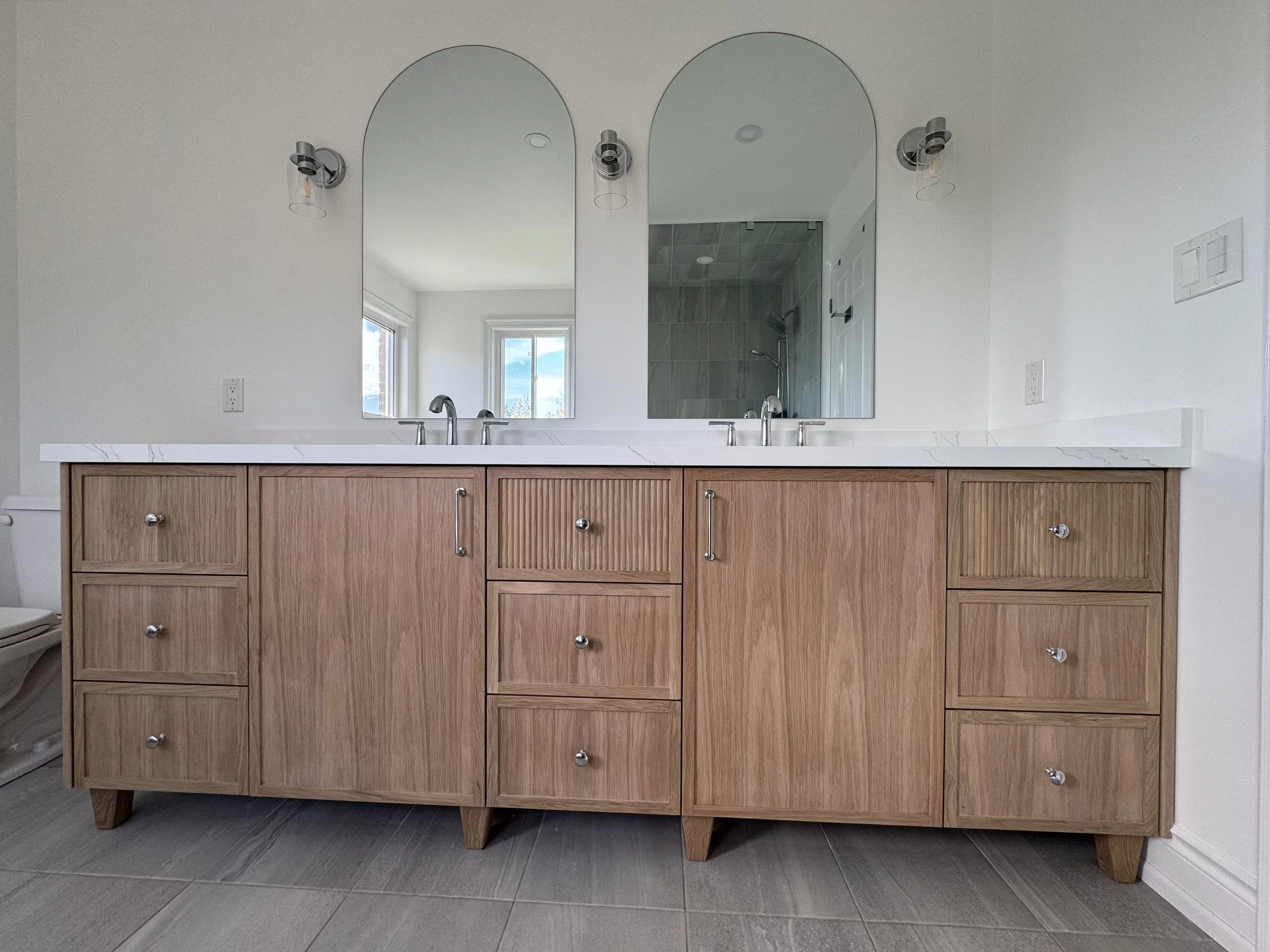 Double vanity bathroom with custom wooden cabinets, white marble countertop, two mounted mirrors, and two wall-mounted lights.