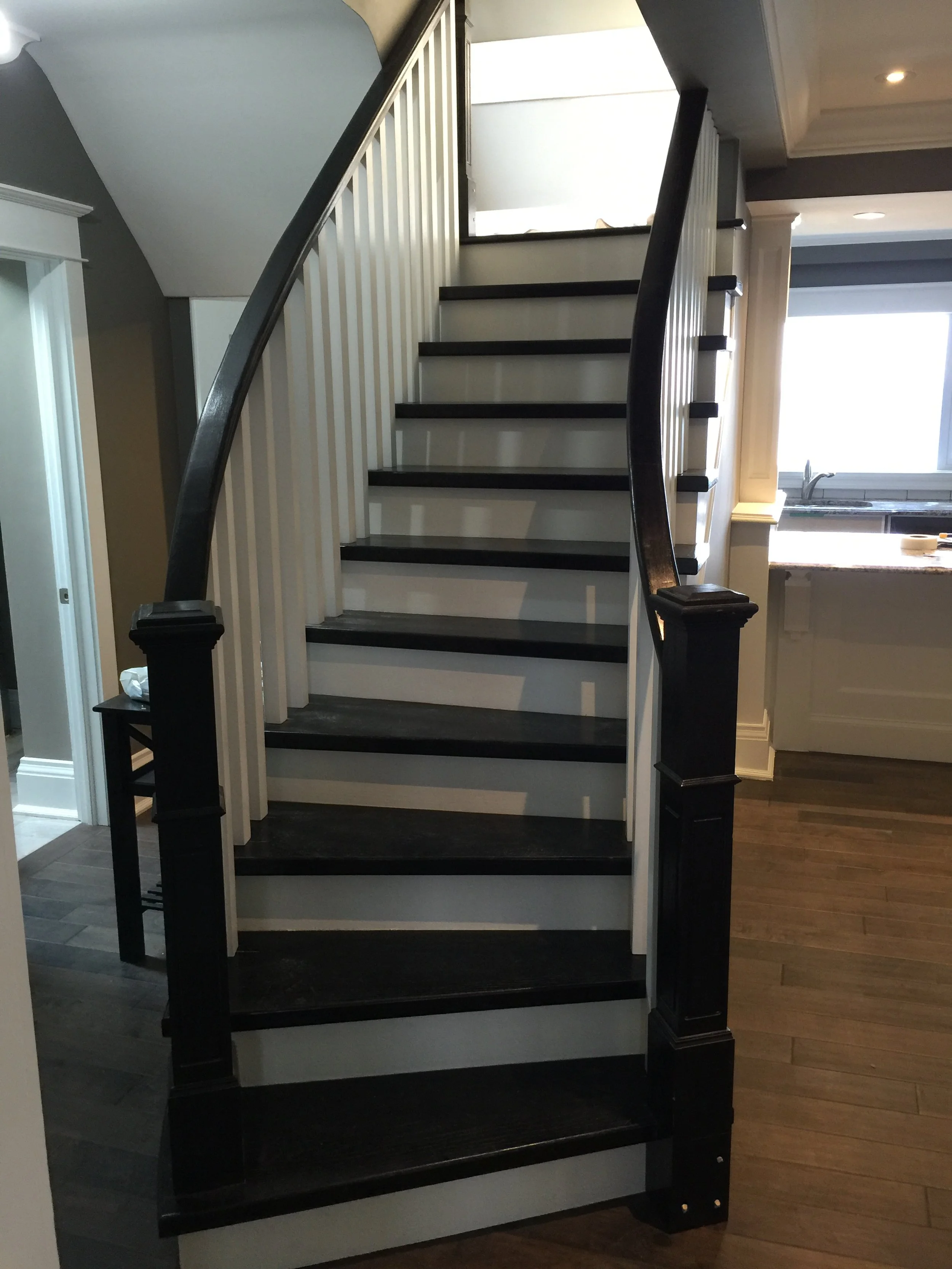 Interior view of a staircase with black treads and white risers, with a dark wooden handrail and white vertical balusters, leading up to an open doorway at the top.