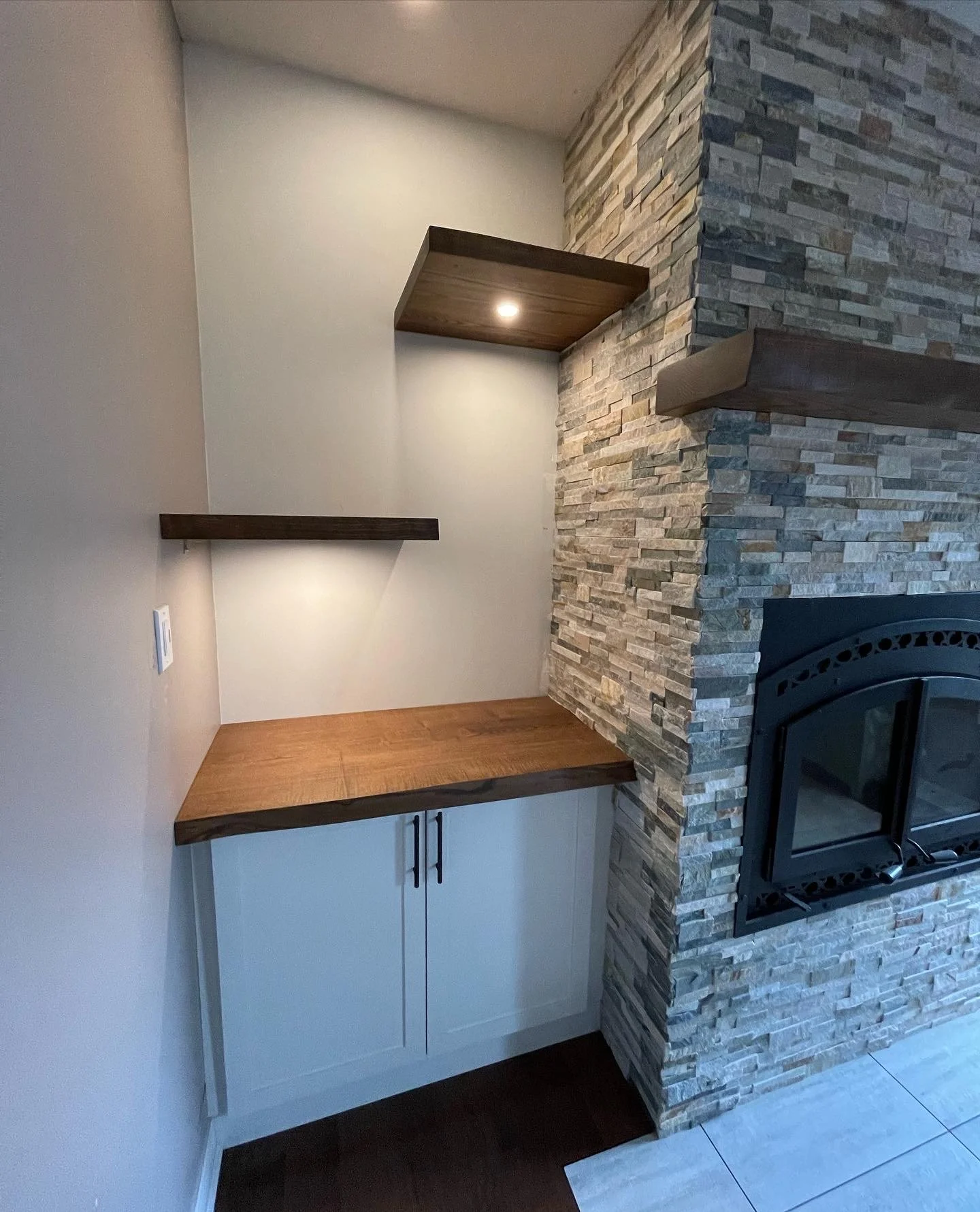 Corner of a room with a built-in cabinet, wooden countertop, and floating shelf, adjacent to a fireplace with a stone facade.
