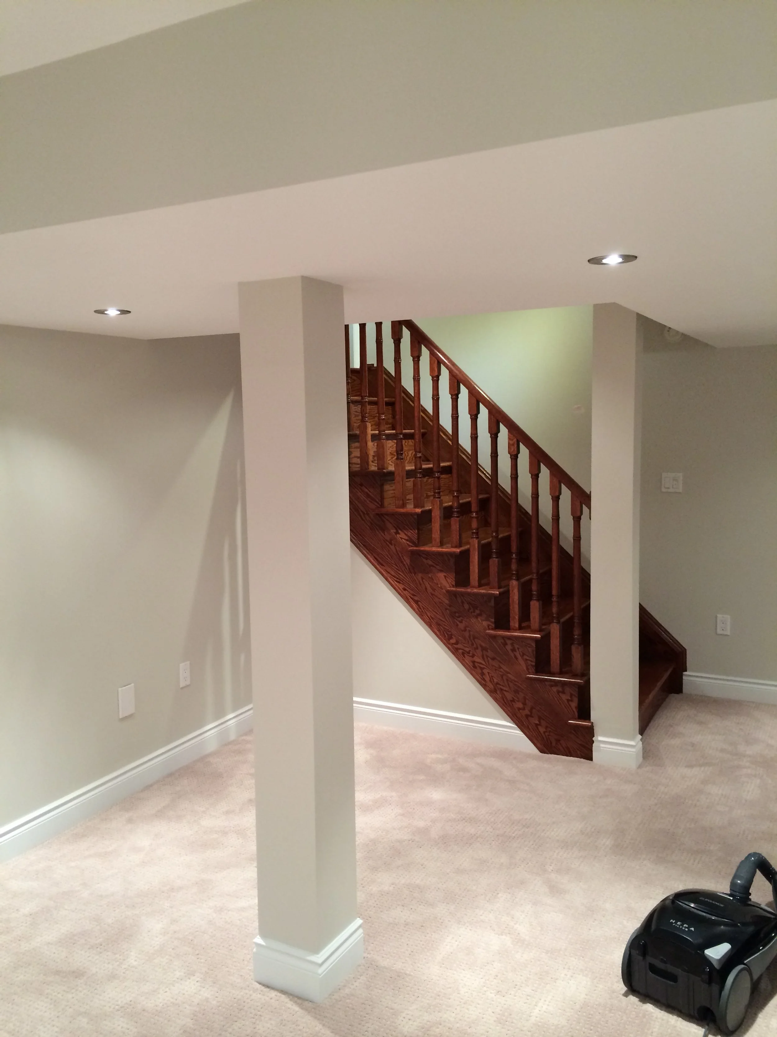Empty basement renovation with beige carpet, white walls, ceiling with recessed lighting, and a wooden staircase.