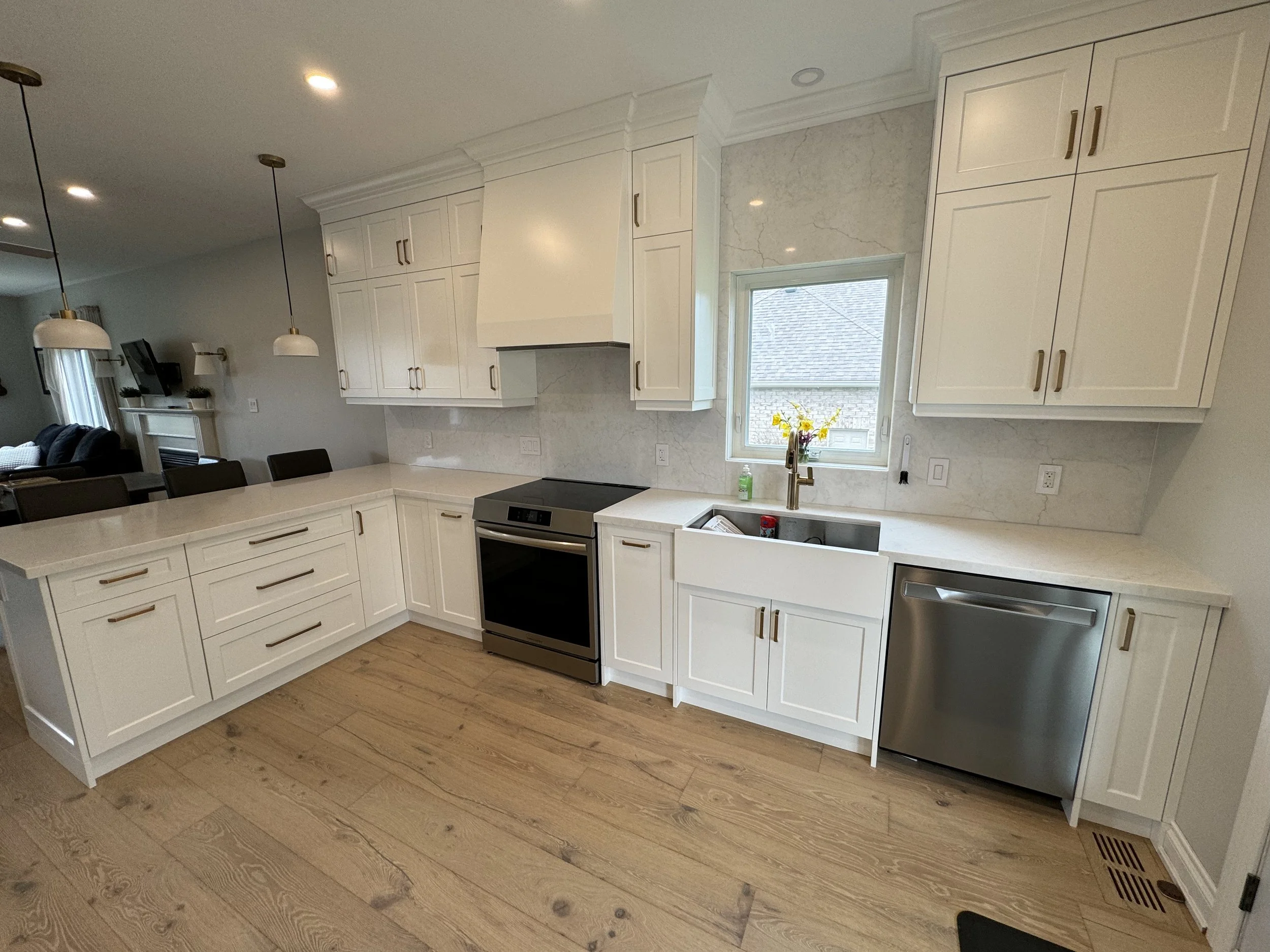 Modern kitchen with white cabinets, marble backsplash, stainless steel stove and dishwasher, farmhouse sink, and gold hardware, hardwood flooring, window above the sink with a view of the roof of a house outside.