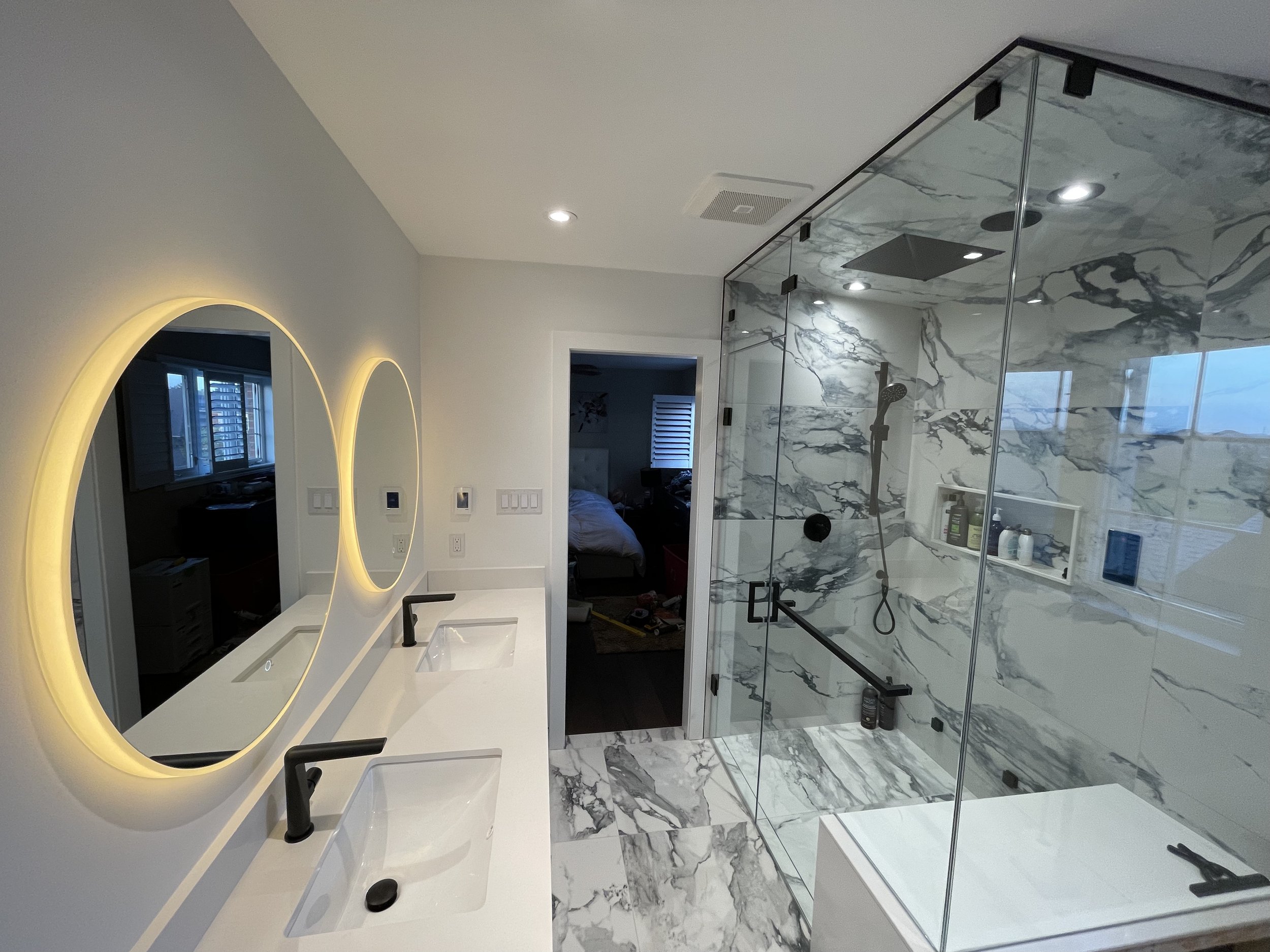 Modern bathroom with dual white sinks, black faucets, illuminated round mirrors, marble shower with glass enclosure, built-in shelf, and a view into a bedroom.