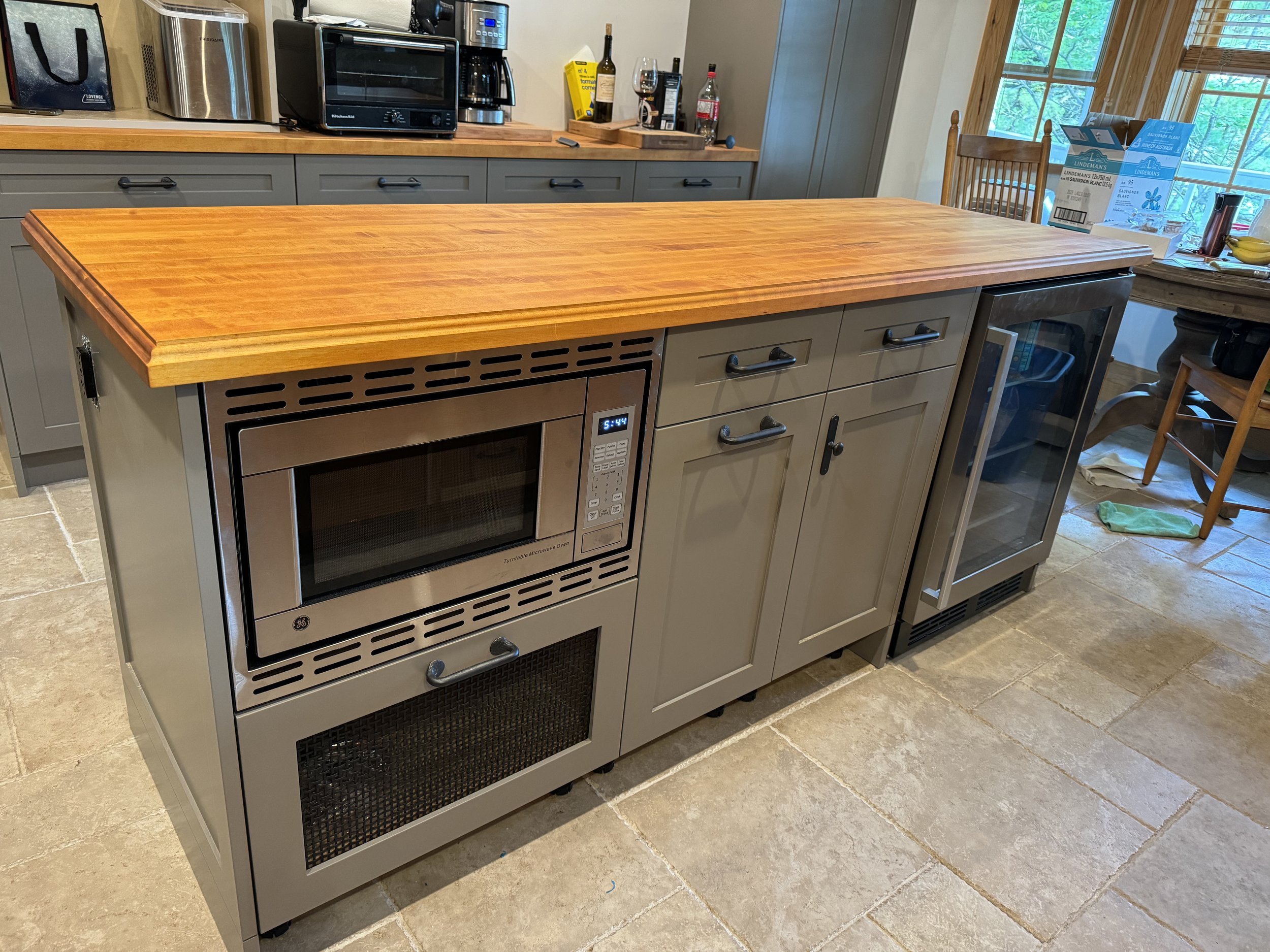 Custom kitchen island with a microwave, cabinets, a wine fridge, and a wooden countertop in a kitchen with tiled floor and windows.