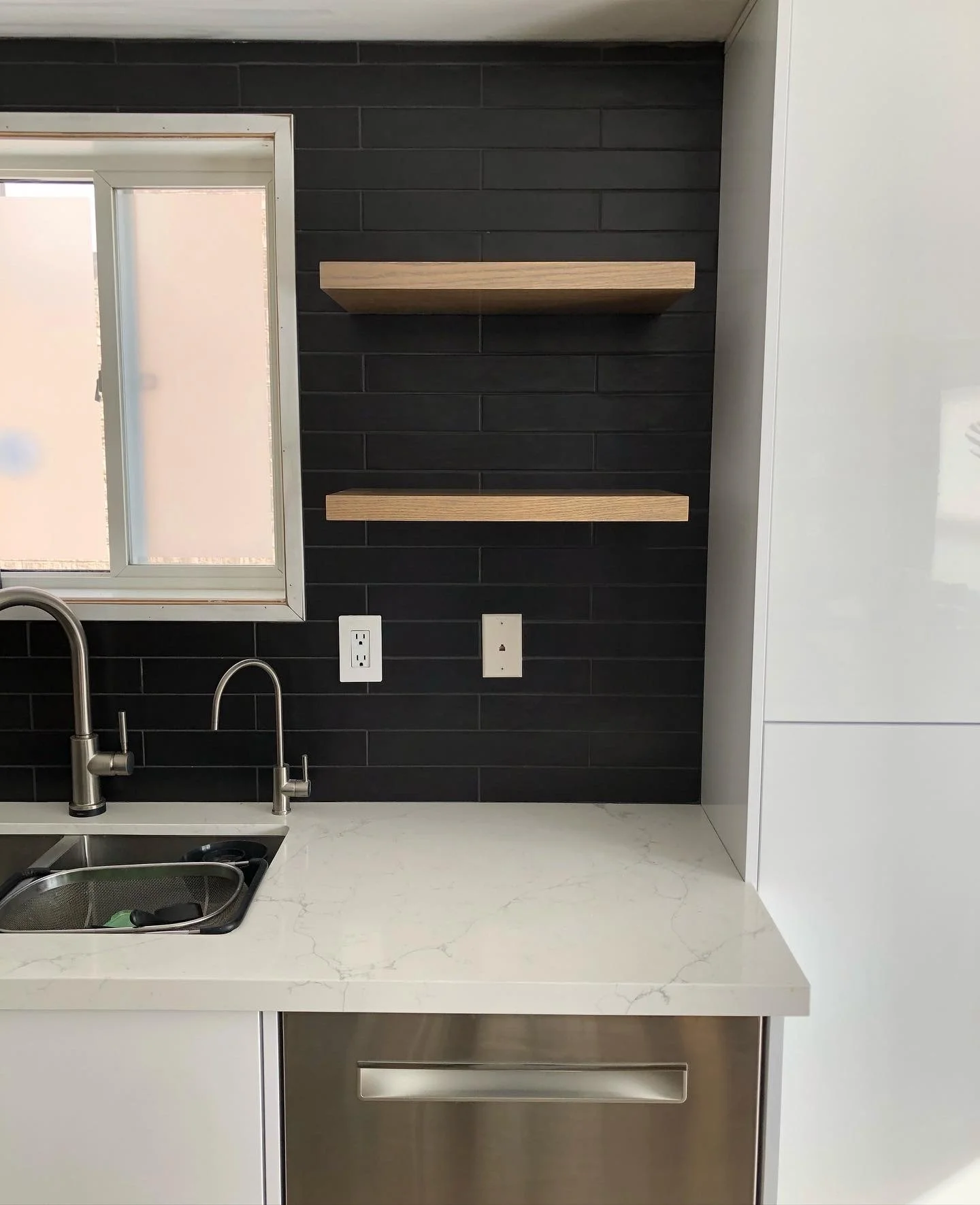 Modern South Georgian Bay custom kitchen with white countertop, stainless steel sink, black subway tile backsplash, two wooden shelves on black wall, and a window with frosted glass.